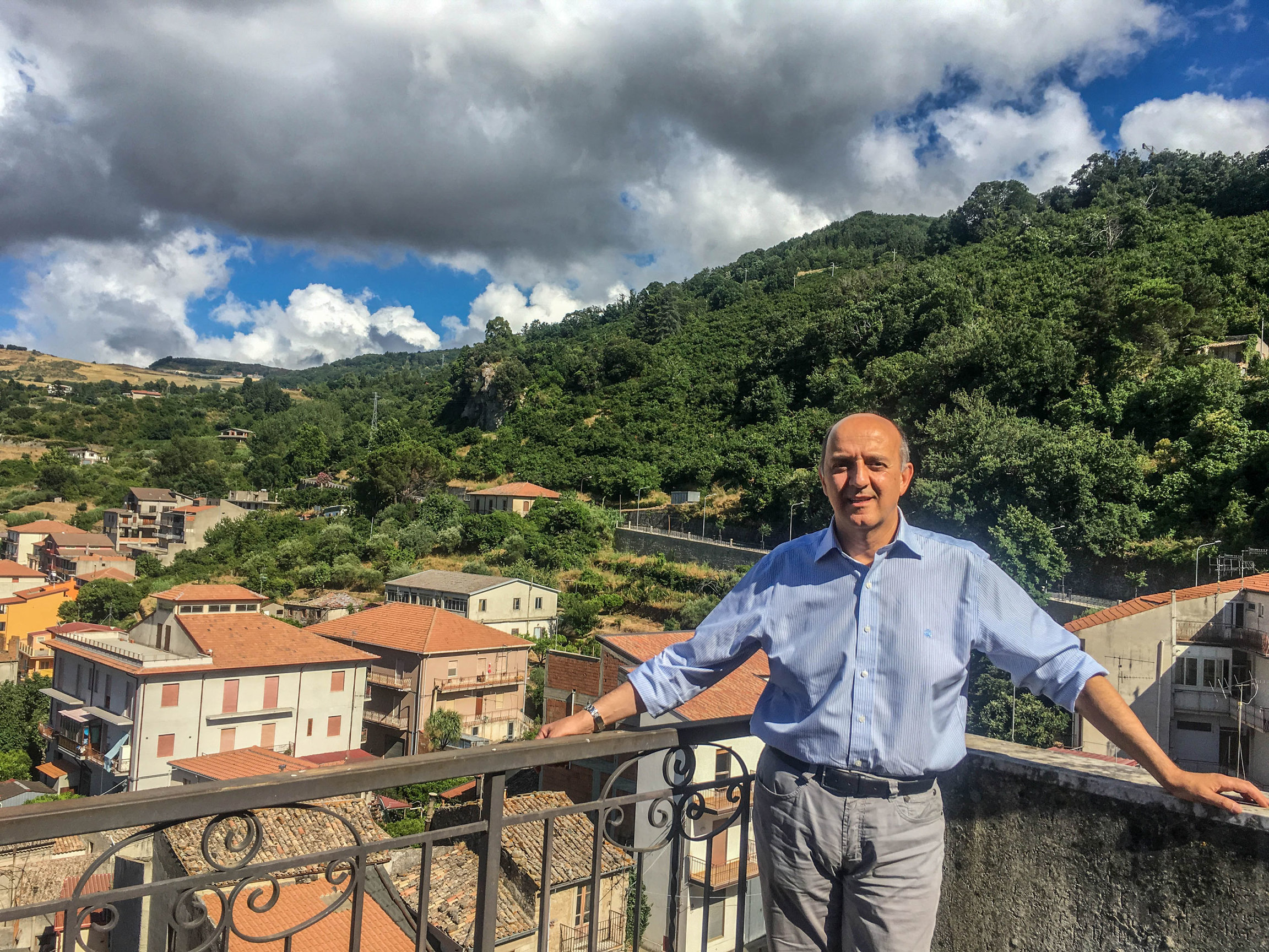 Salvatore Fiore, the mayor of San Piero Patti, Italy, said his town had watched the success of offering homes for sale for 1 euro in other places and wanted to try it out here. A man is shown standing on a balcony wearing a white button-down shirt with a lush green hillside in the background.