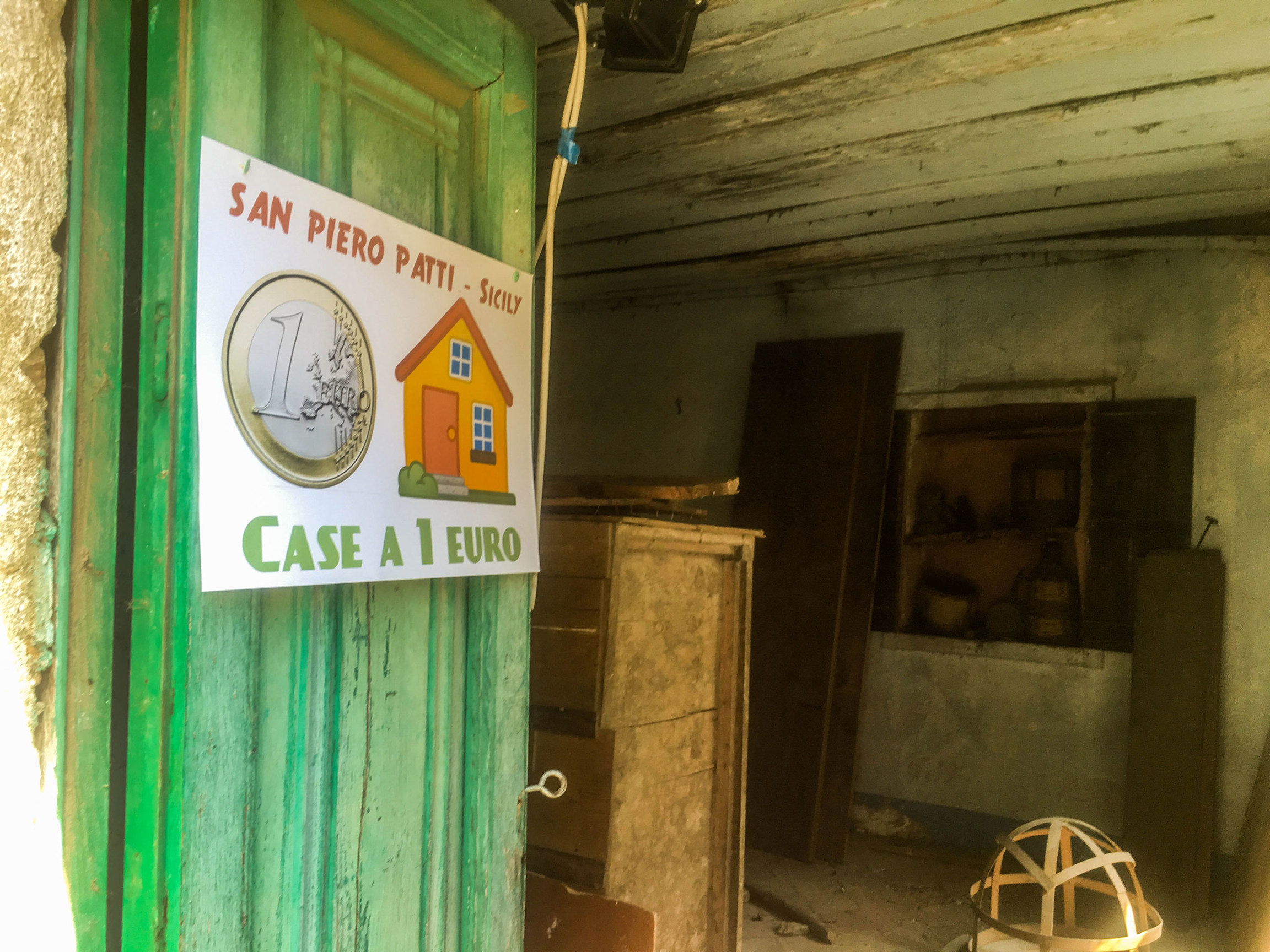 Maria Concetta Abramo's house is on sale for 1 euro — as advertised by the sign on the door. A green door is shown ajar with a sign posted on it saying "San Piero Patti, Sicly, Case a 1 euro."