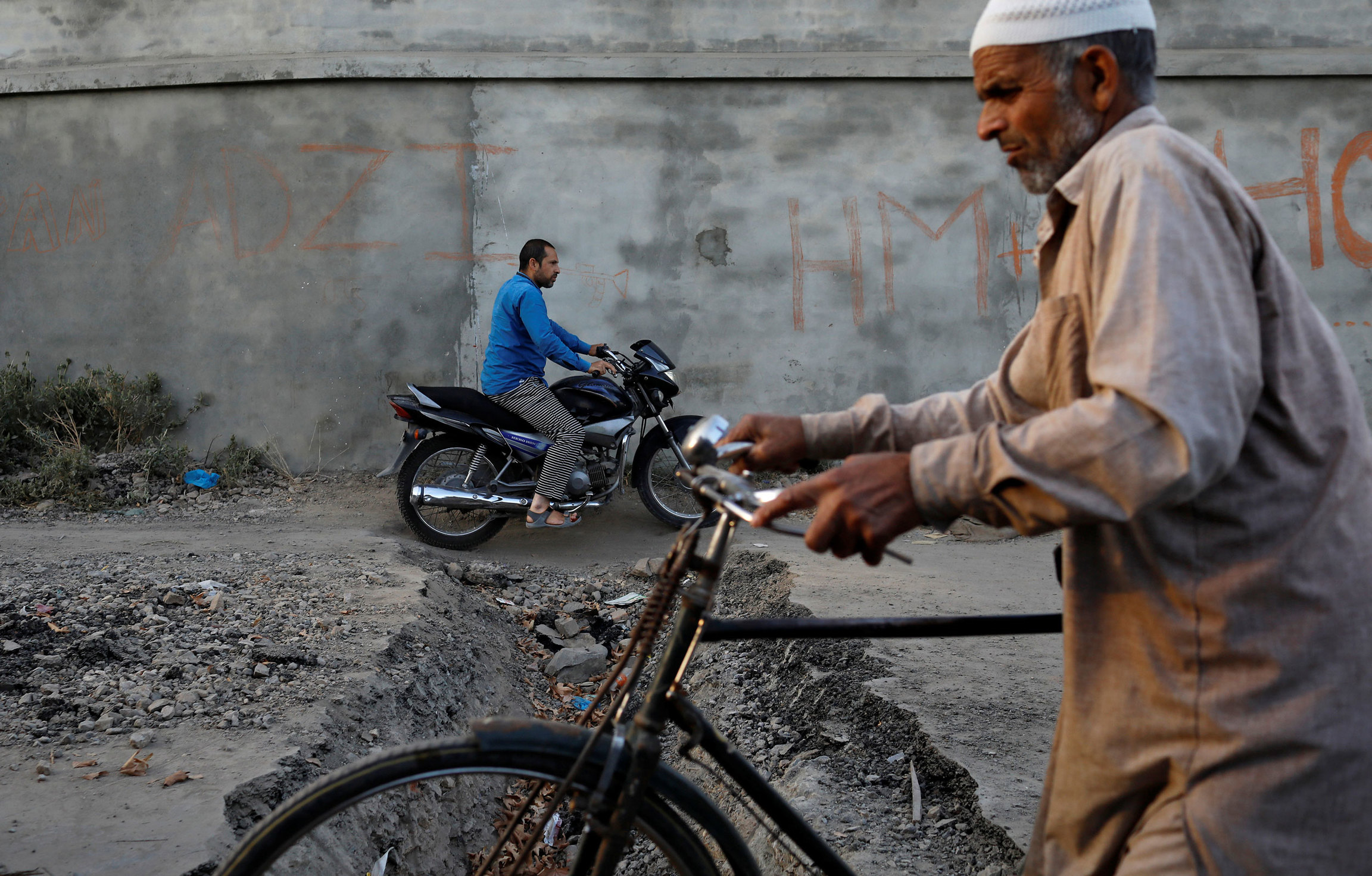 Kashmiris ride past a trench dug by residents to prevent Indian security forces from entering Anchar neighborhood. Two men are shown passing each other, one riding a motocycle and the other a bicycle.