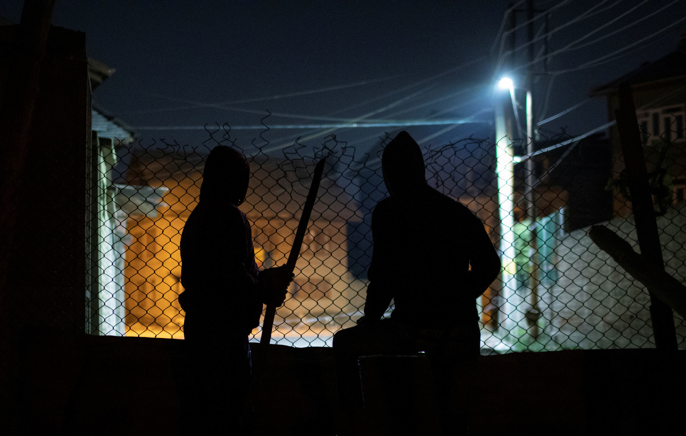 Kashmiri men stand guard at night at a barricade to prevent Indian security forces from entering Anchar neighborhood. Two men are shown in shadow with a chain link fence in front of them with only the light of a streetlamp.
