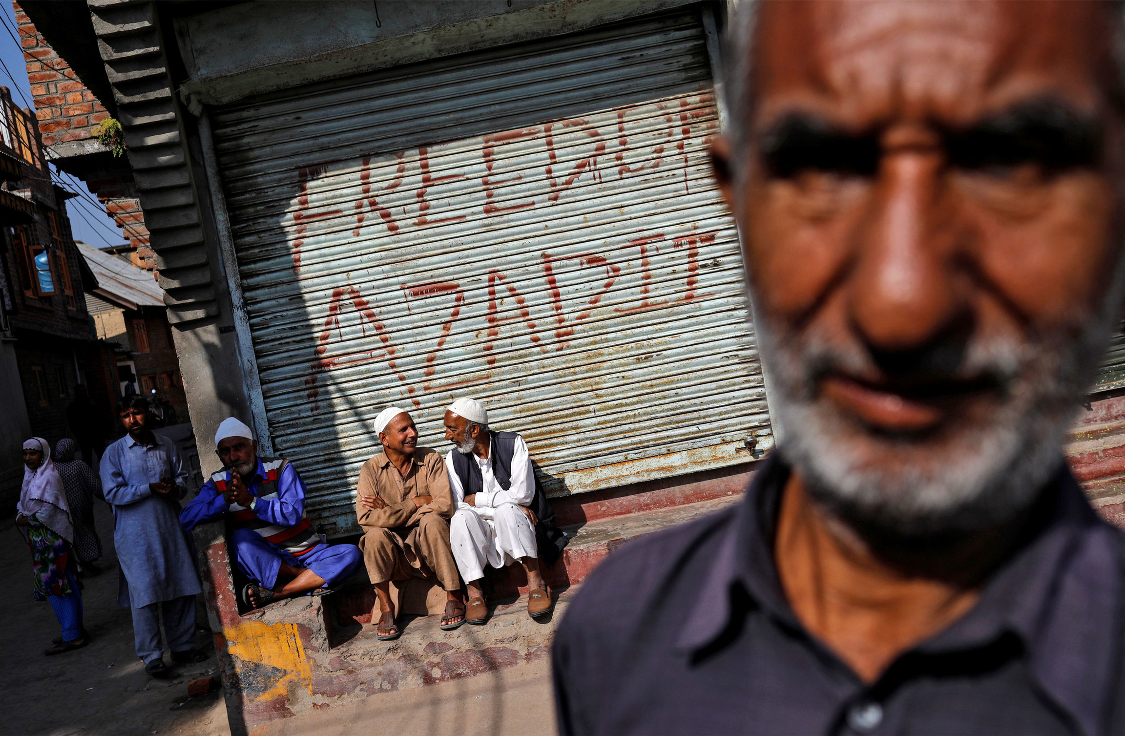 Kashmiri men sit in front of pro-independence graffiti sprayed on the shutter of a closed shop. A man is shown in the near ground out of focus with a row of men shown sitting on a step in the background.