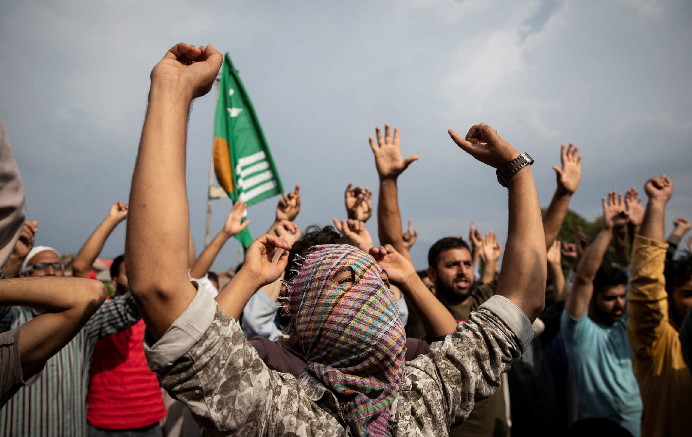 Kashmiris shout slogans in Anchar neighborhood after Friday prayers. A group of people are shown, many with their hands in the air and a man in the center wearing a face covering.