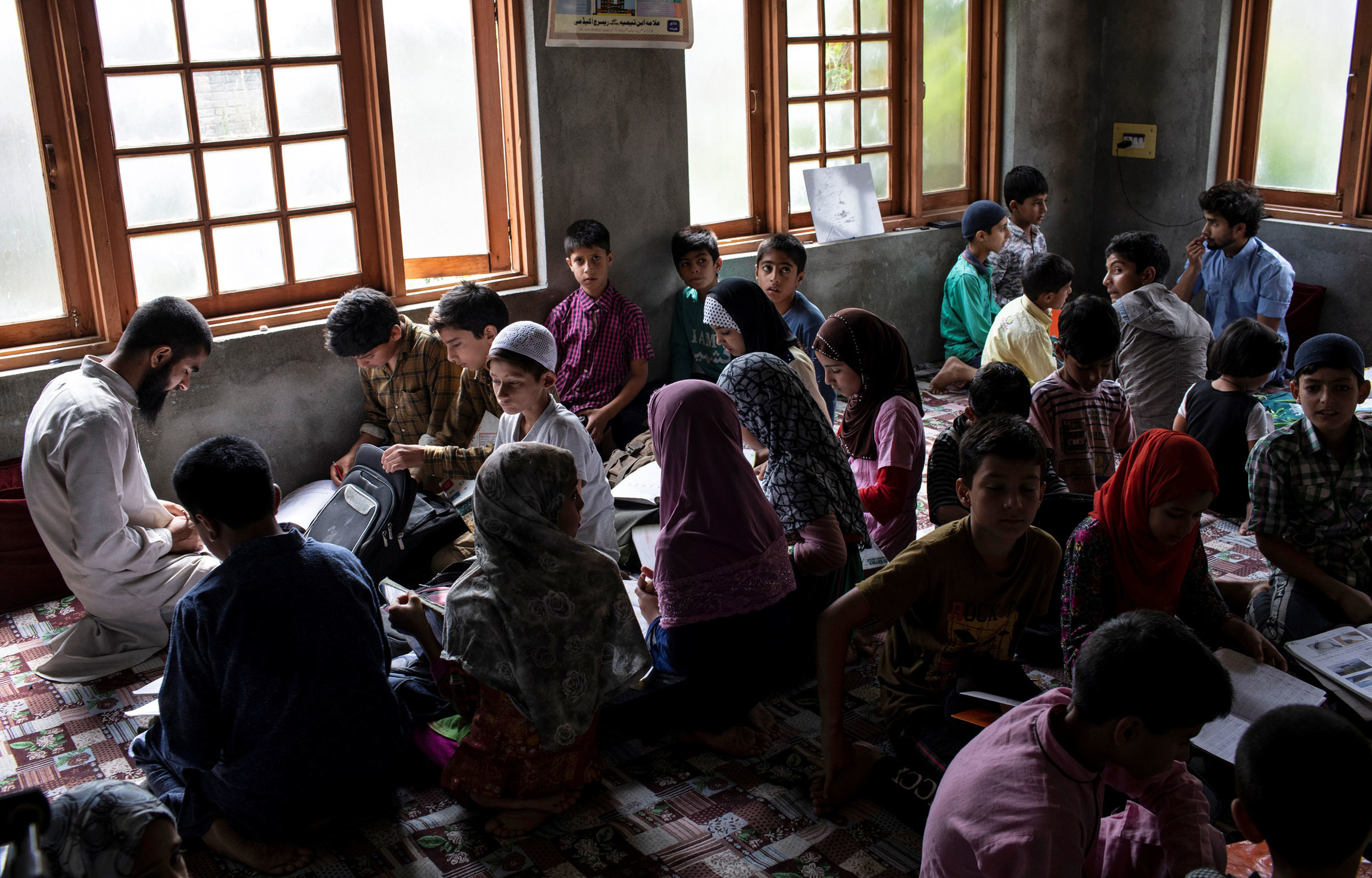 Kashmiri college students teach children inside a house. A classroom of children are shown sitting on the floor and doing school work.