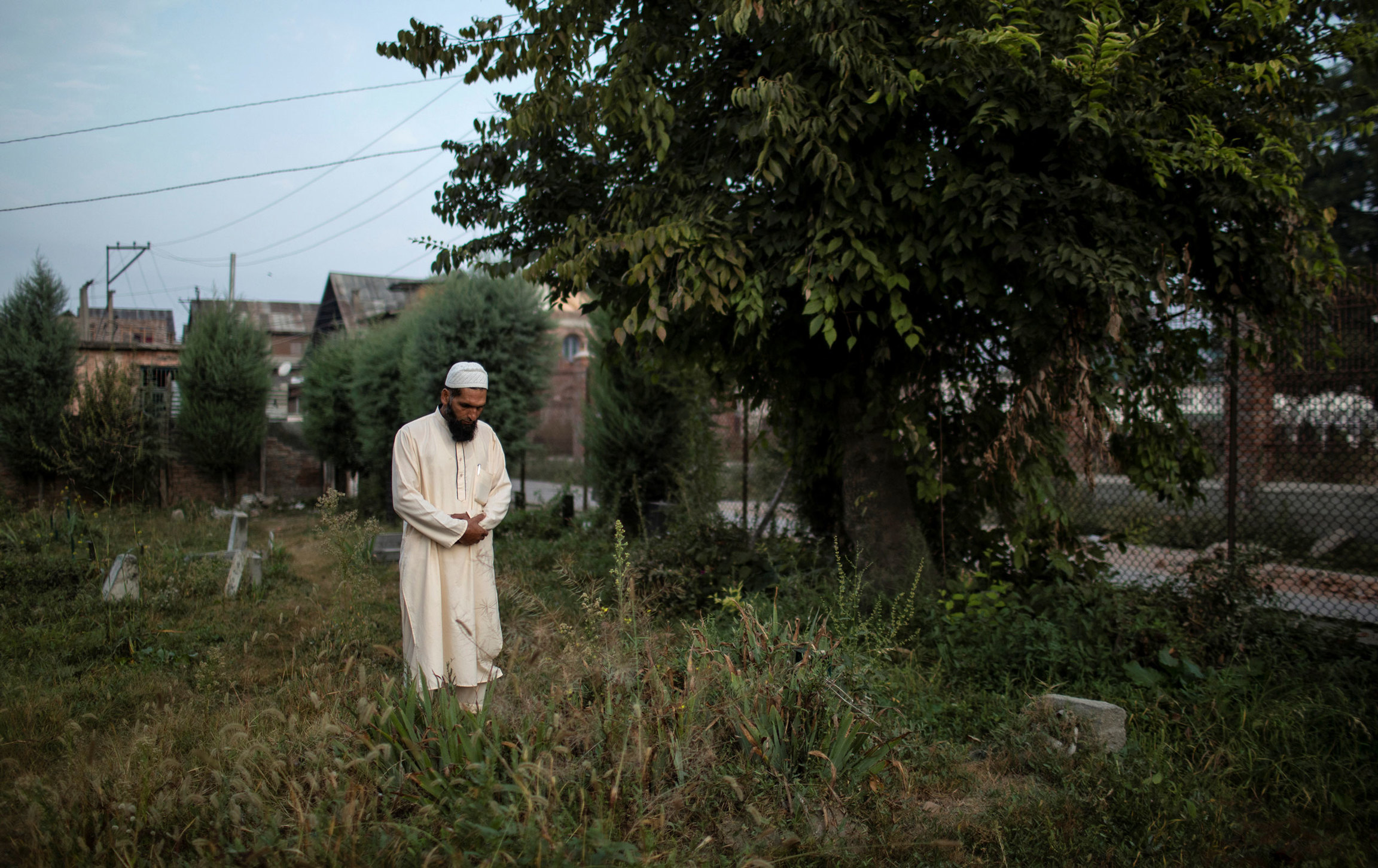 A Kashmiri man prays at a graveyard. A man is shown wearing all white and a white cap with his hands clasped in prayer.