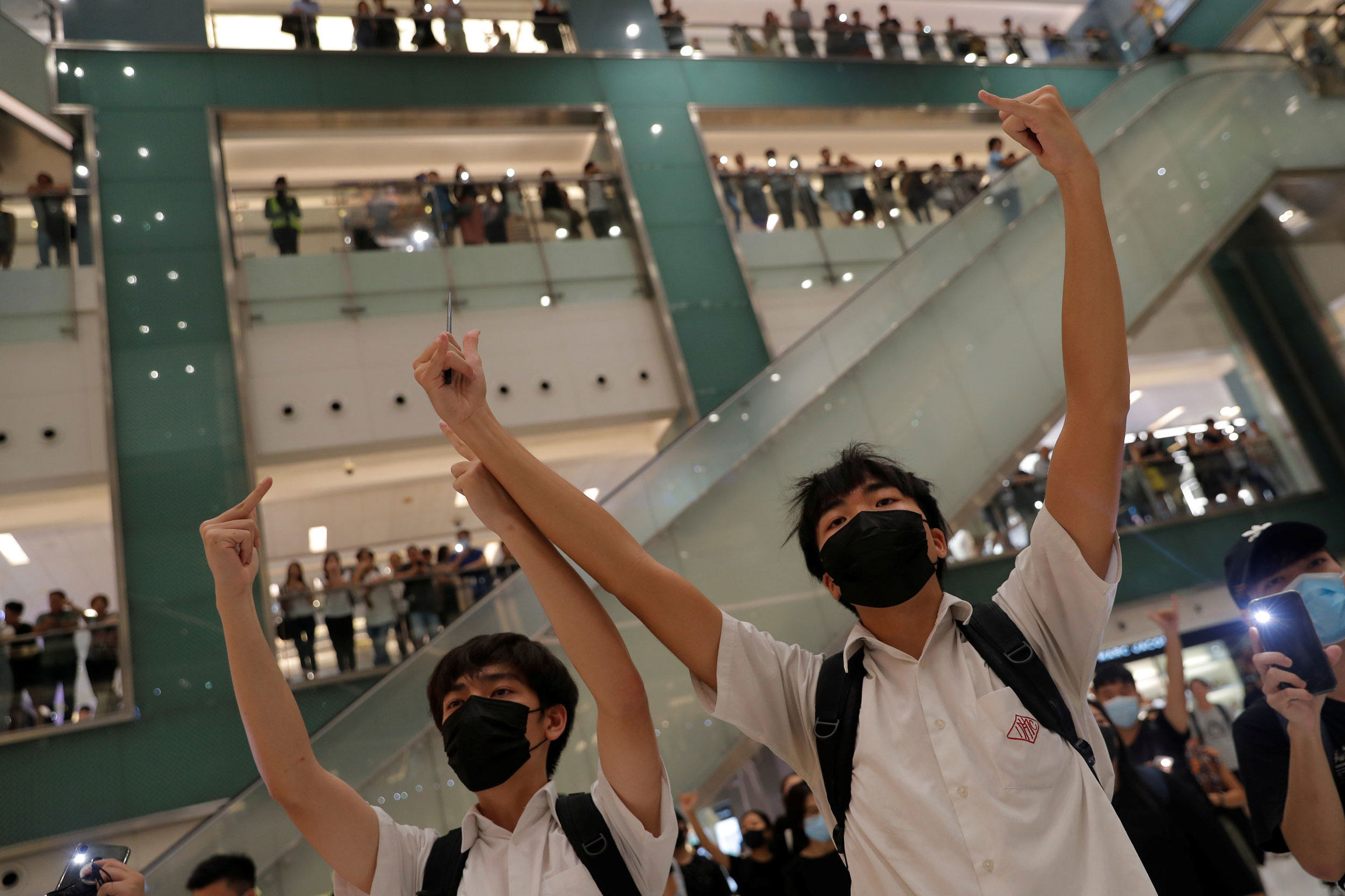 Anti-government protesters gather inside a mall in Sha Tin, Hong Kong, Oct. 3, 2019. Two men are shown wearing back packs and black masks with their arms raised in the air gesturing.