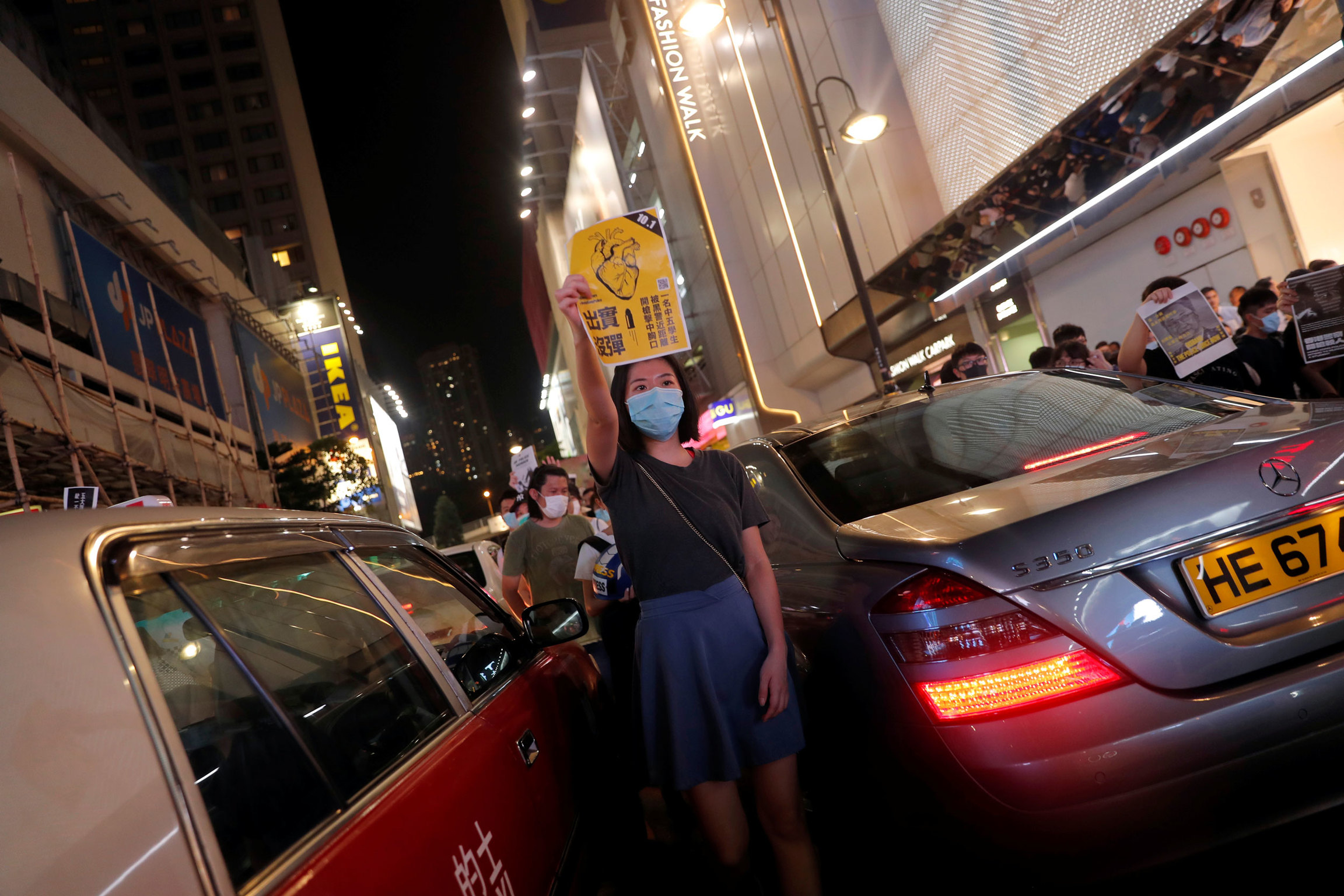 Anti-government protesters block a street during a march in Causeway Bay in Hong Kong. A woman is shown wearing a hospital mask and standing inbetween two cars.
