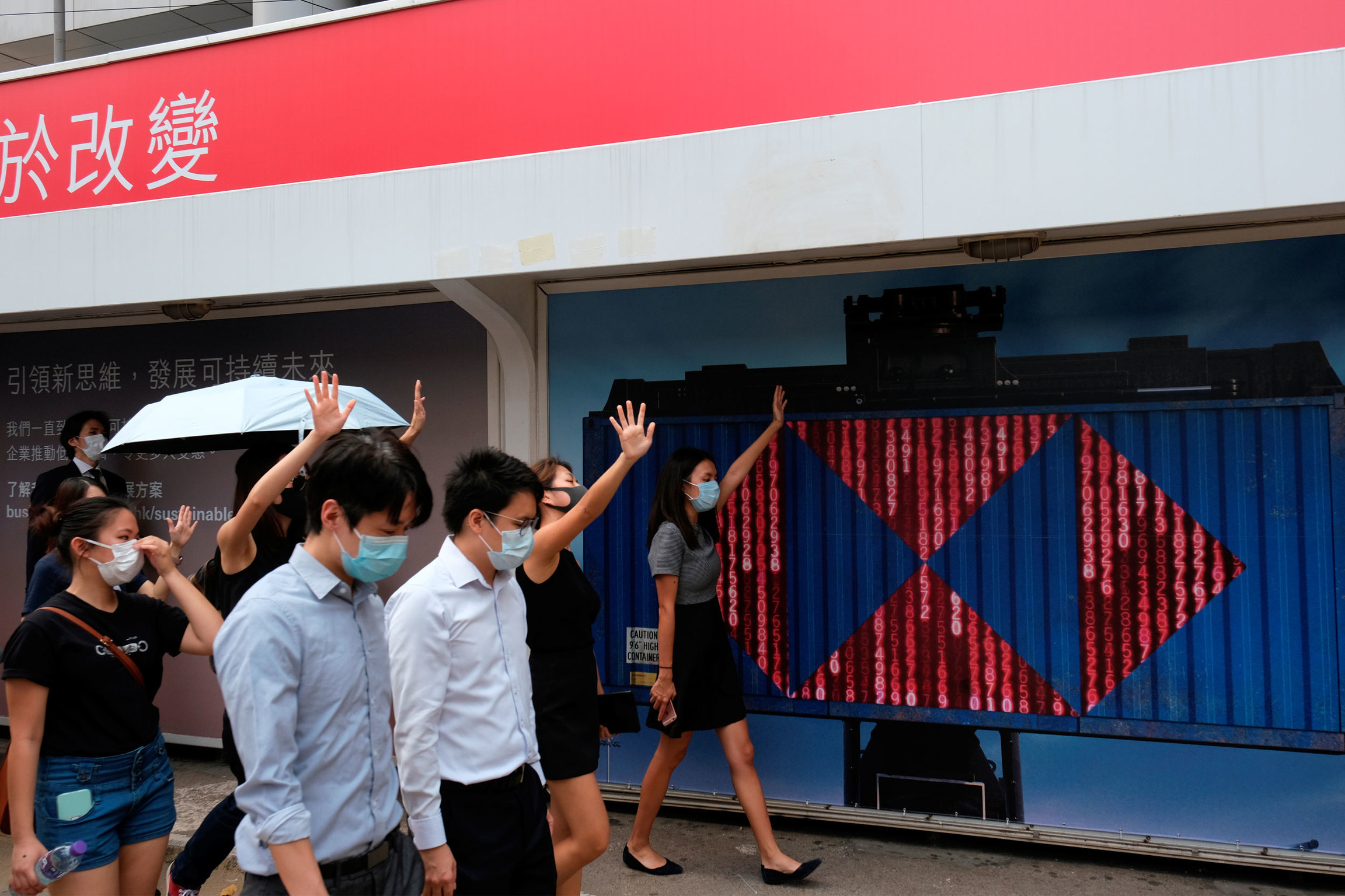 Anti-government demonstrators walk past a board with the logo of HSBC during a flash mob protest in Hong Kong. A small crowd of people are shown walking and all are wearing face masks and one person is carrying an umbrella.