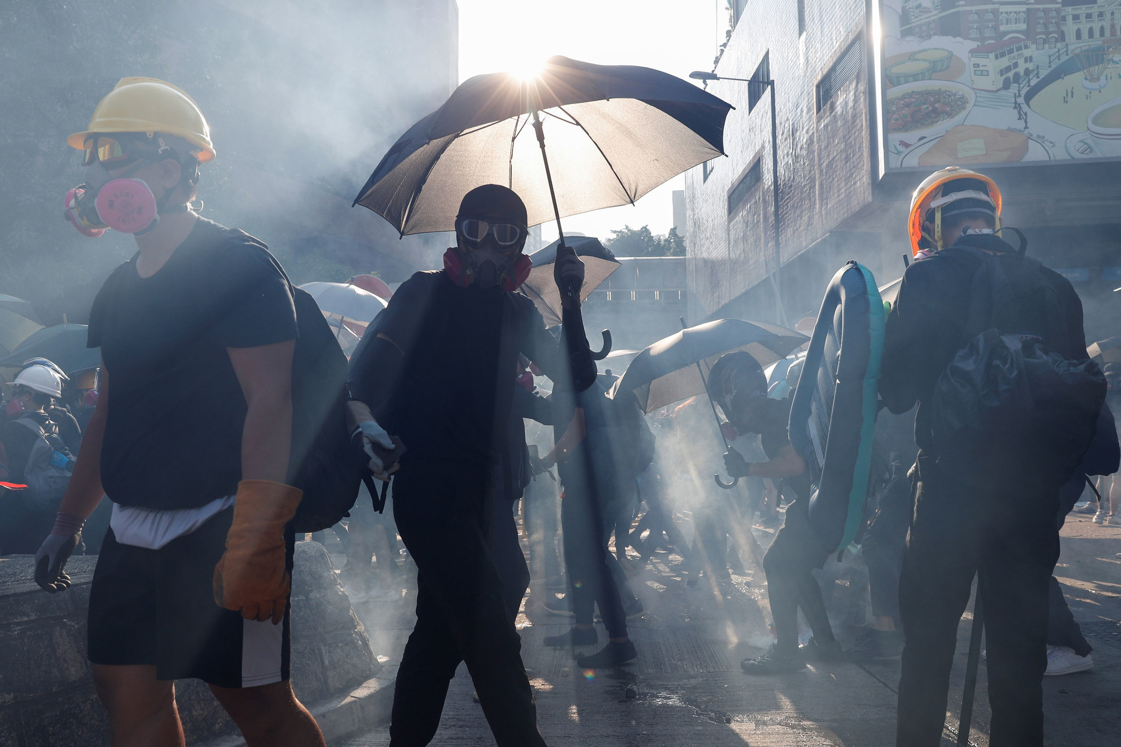 Anti-government protesters react in a cloud of tear gas during a demonstration on China's National Day. Several protesters are shown wearing gas masks and one holding an umbrella with the sun glaring above it.