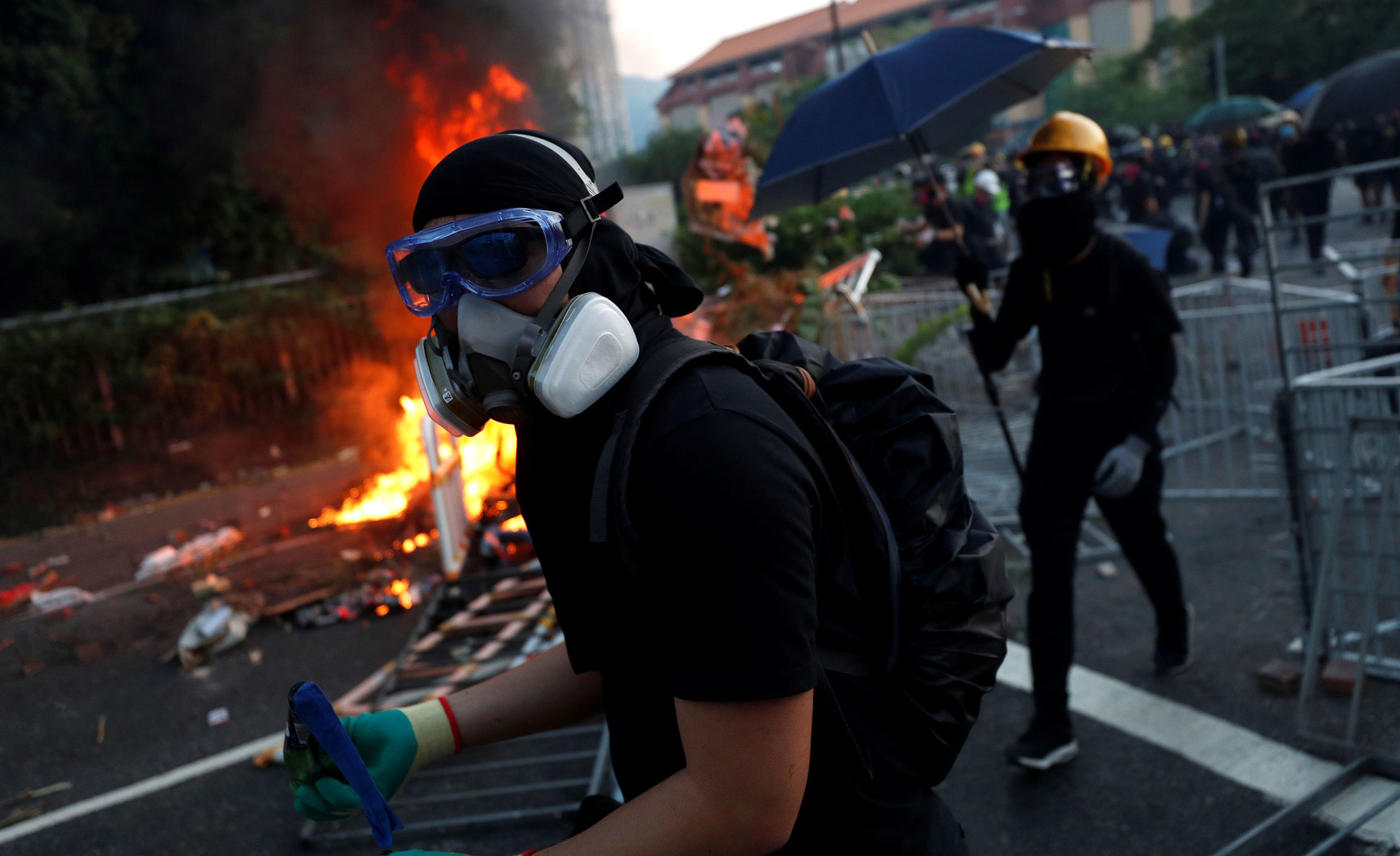 An anti-government protester walks past a fire during a demonstration in Sha Tin district in Hong Kong. A protester is shown wearing goggles and a gas mask walking past a blazing fire.