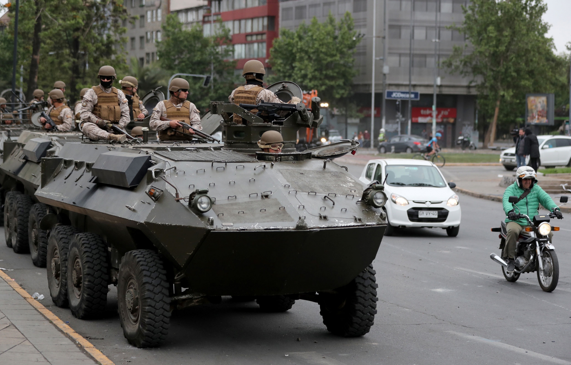 Soldiers ride an armored transporter as they patrol the Santiago, Chile, after widespread protests, Oct. 20, 2019. A line of armored transporter trucks are shown with armed soldiers riding on top and a man on a motorcycle passing by.