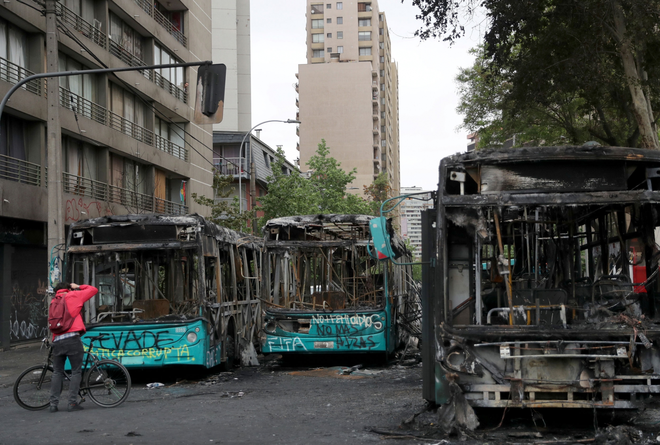 A man takes photos of burned buses, which were destroyed duirng a previous day's protest in Santiago, Chile, Oct. 20, 2019. A man is shown with a bicycle standing in front of three totally burned out buses in a street.
