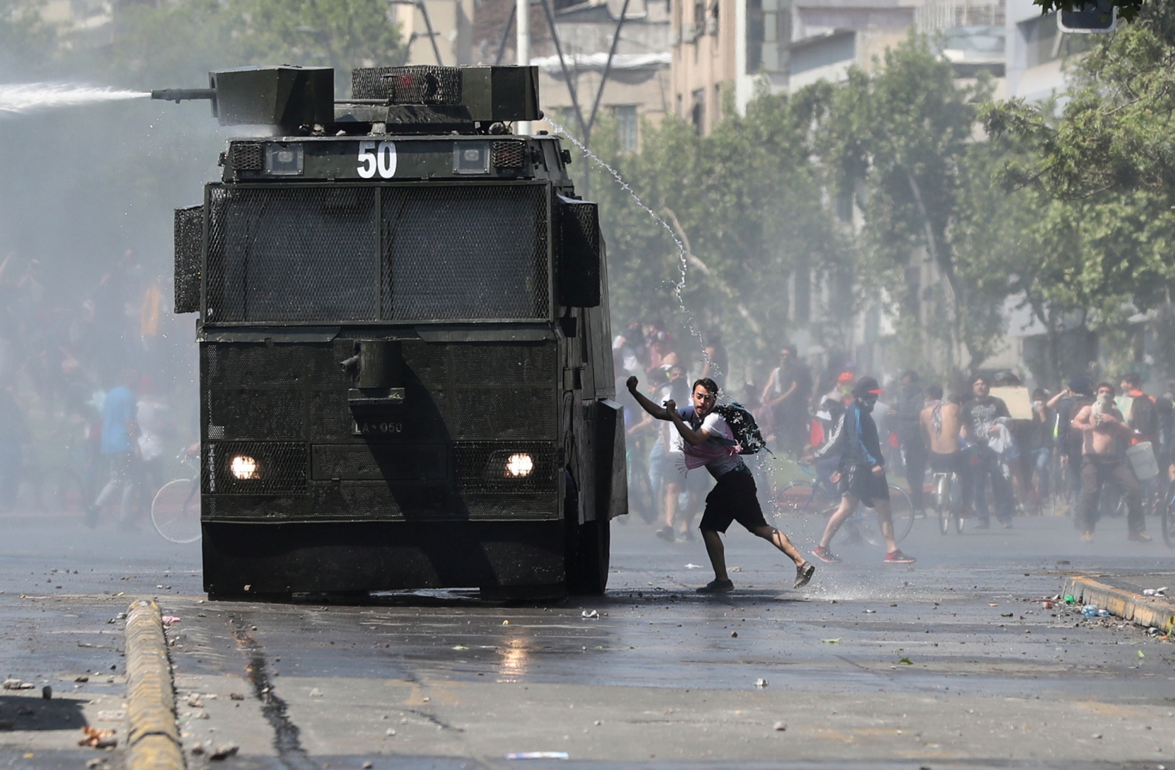 A demonstrator approaches a riot police water cannon during a protest in Santiago, Chile. A man is shown wearing shorts and a t-shirt while getting sprayed by a police water cannon from above.