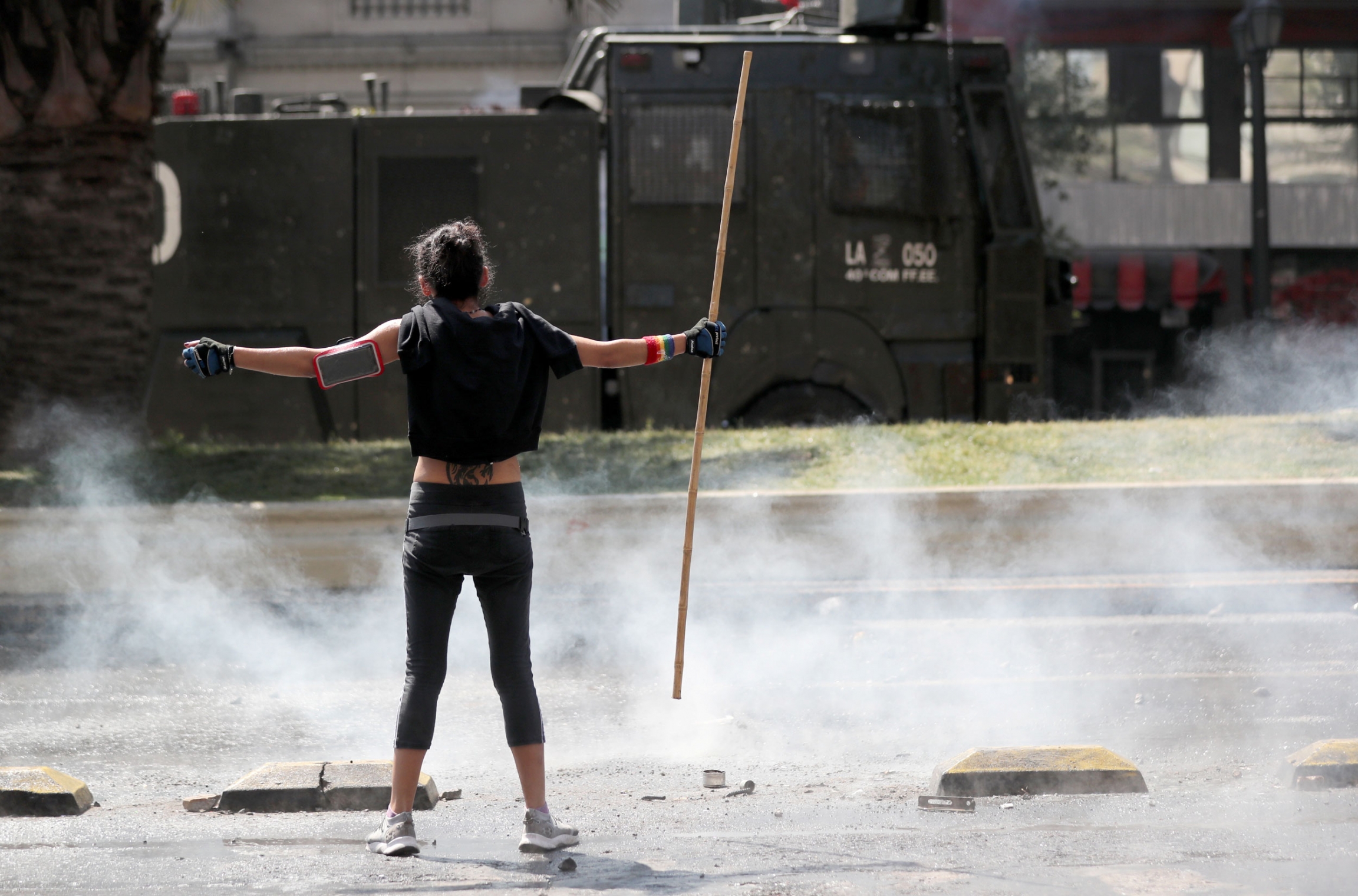 A woman stands with her arms out during a protest against Chile's state economic model in Santiago, Oct. 20, 2019. A woman is shown stands with her arms out, holding a stick, with smoke around her and a military truck in the distance.