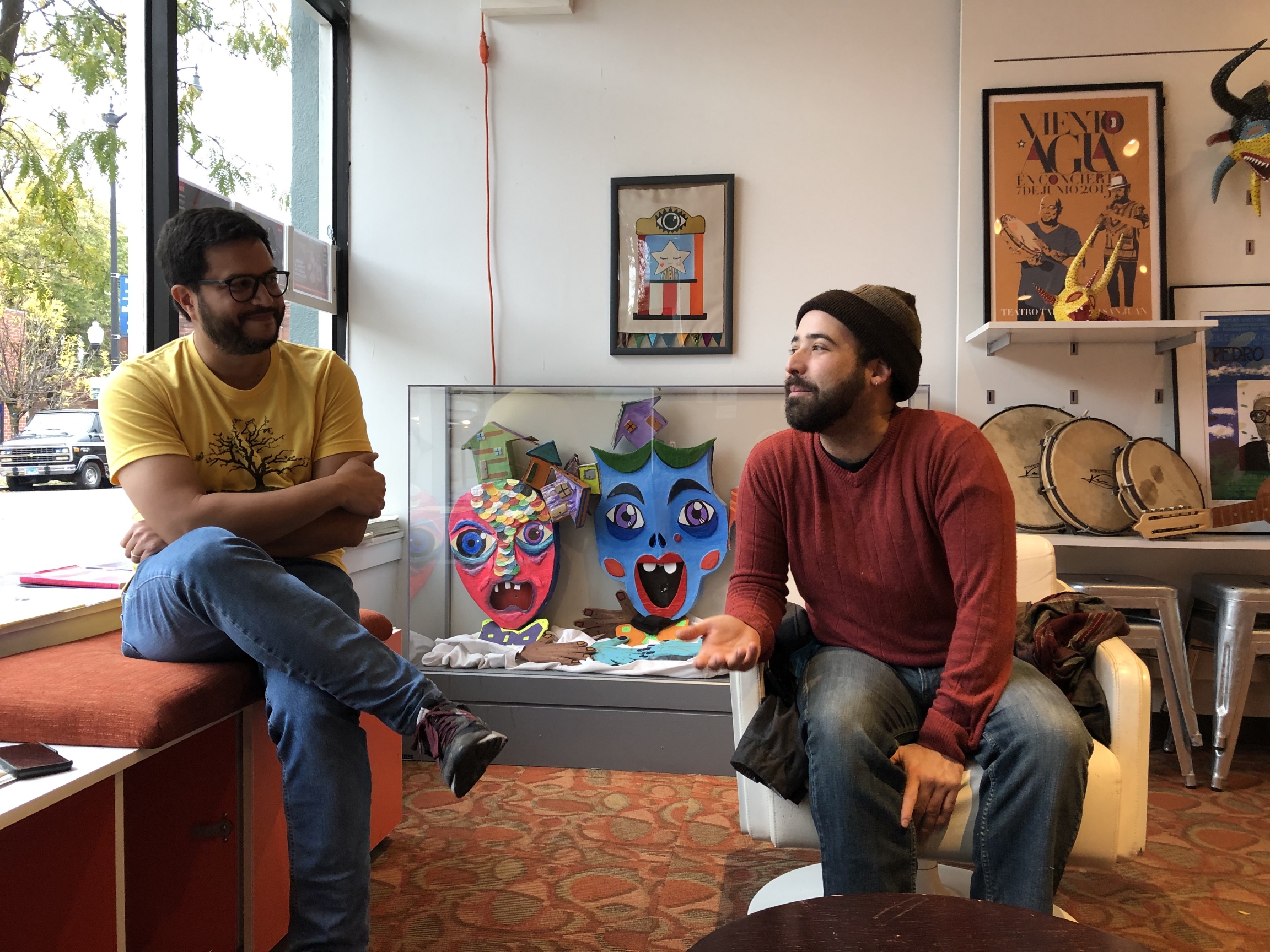 Omar Torres Kortright, executive director of the Segundo Ruiz Belvis Cultural Center (left) sits with actvist Miguel Alvelo (right) in the lobby of SRBCC, Chicago, Illinois, Oct. 21, 2019. Two men sit in a sun-lit lobby with books and plants.