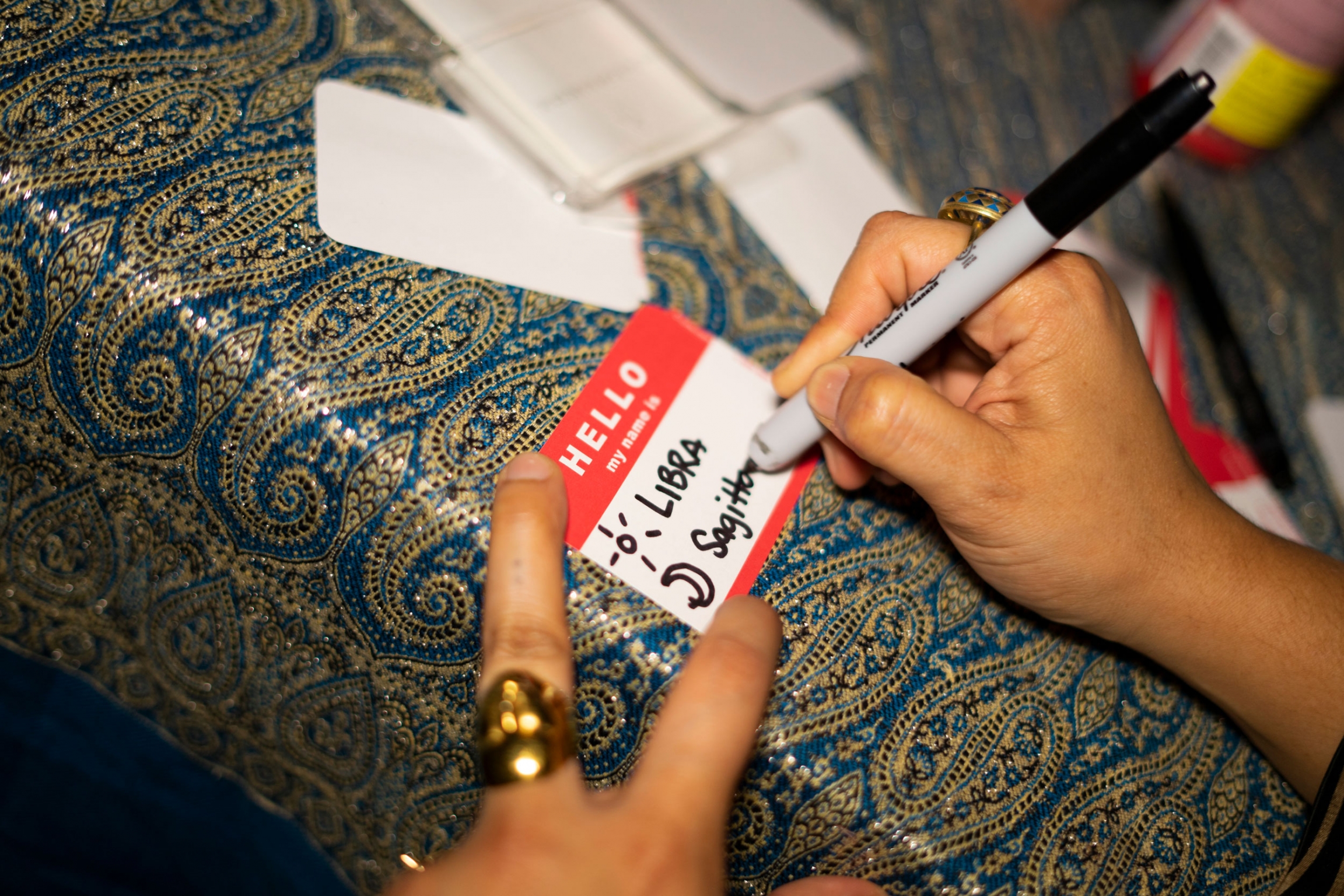 The name tags at an Eleventh House party display astrological signs. A person is writing the word, "Libra" on a name tag.