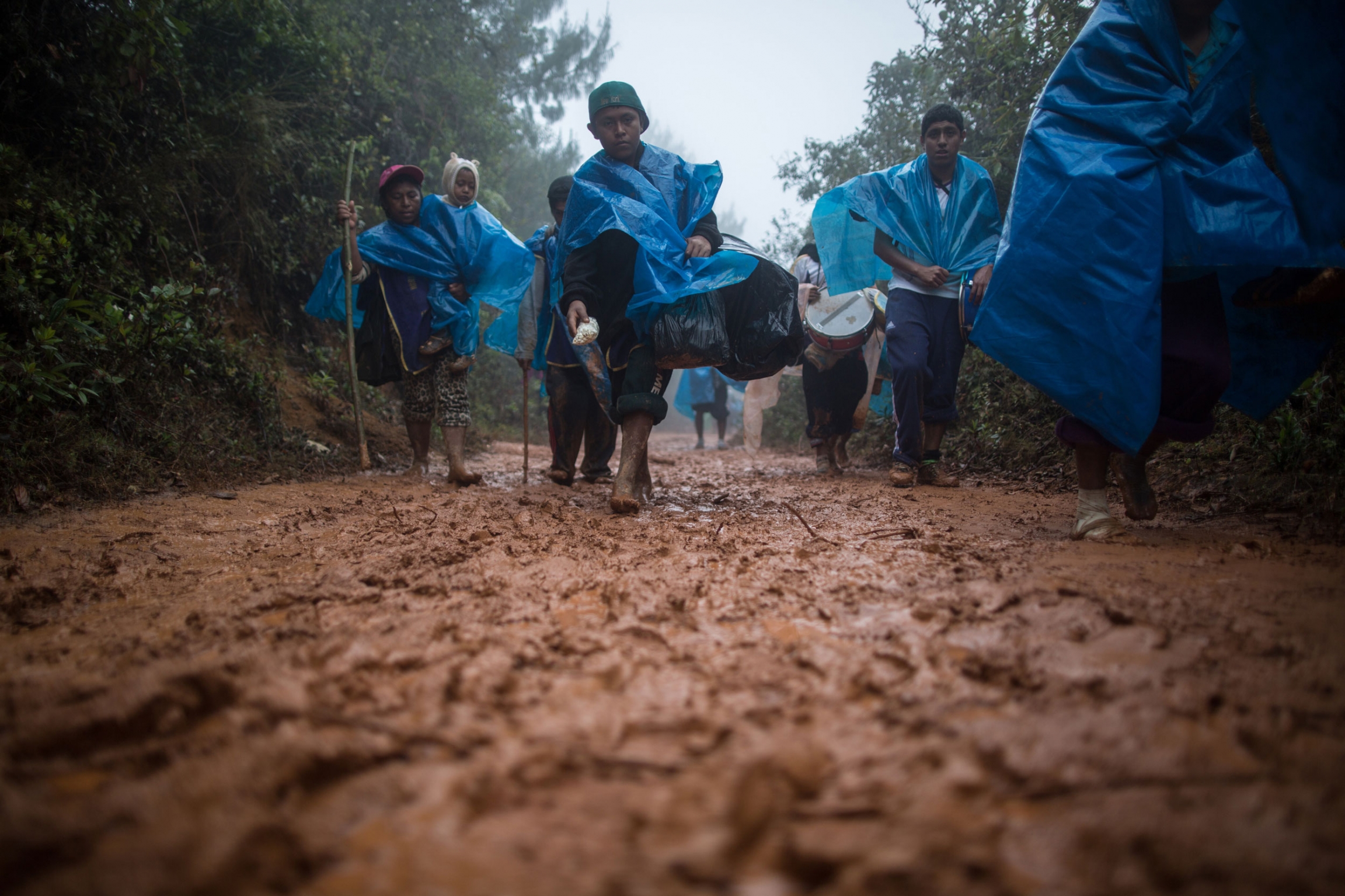 The rains in the Andes can make the trip longer and more dangerous. When it rains in the area of the Devil's Nose, the mud makes this last section before reaching Ayabaca, even more difficult. Walking without shoes, as these pilgrims are doing, helps in t Several people are shown walking along a muddy path and wearing blue rain ponchos.