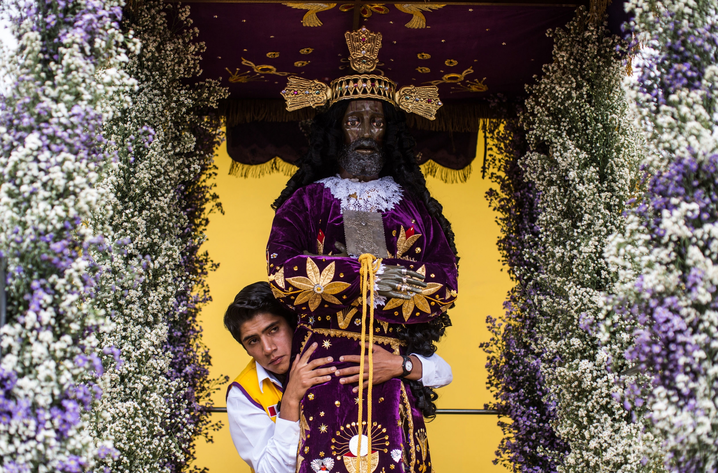 A pilgrim in Ayabaca wraps their arms around a depiction of the Captive. A young man is shown standing behind a carved statue of the Captive which is adorned with a decorative religious robe.
