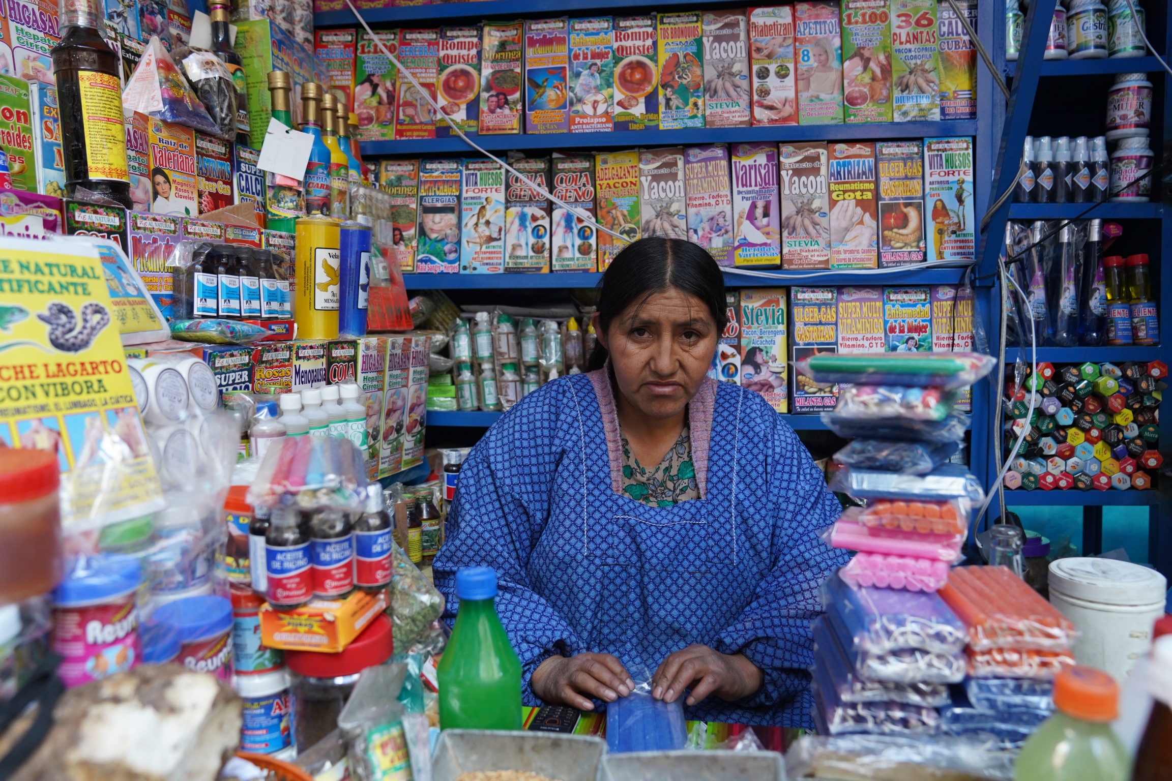 Maria Achu, a traditional medicine merchant, says that she still supports Evo Morales because he has improved the situation of Indigenous people like herself. Her family has benefited from government subsidies. Morales supporter wearing a blue top stands in a fully-stocked shop with bright and colorful products stacked behind her.