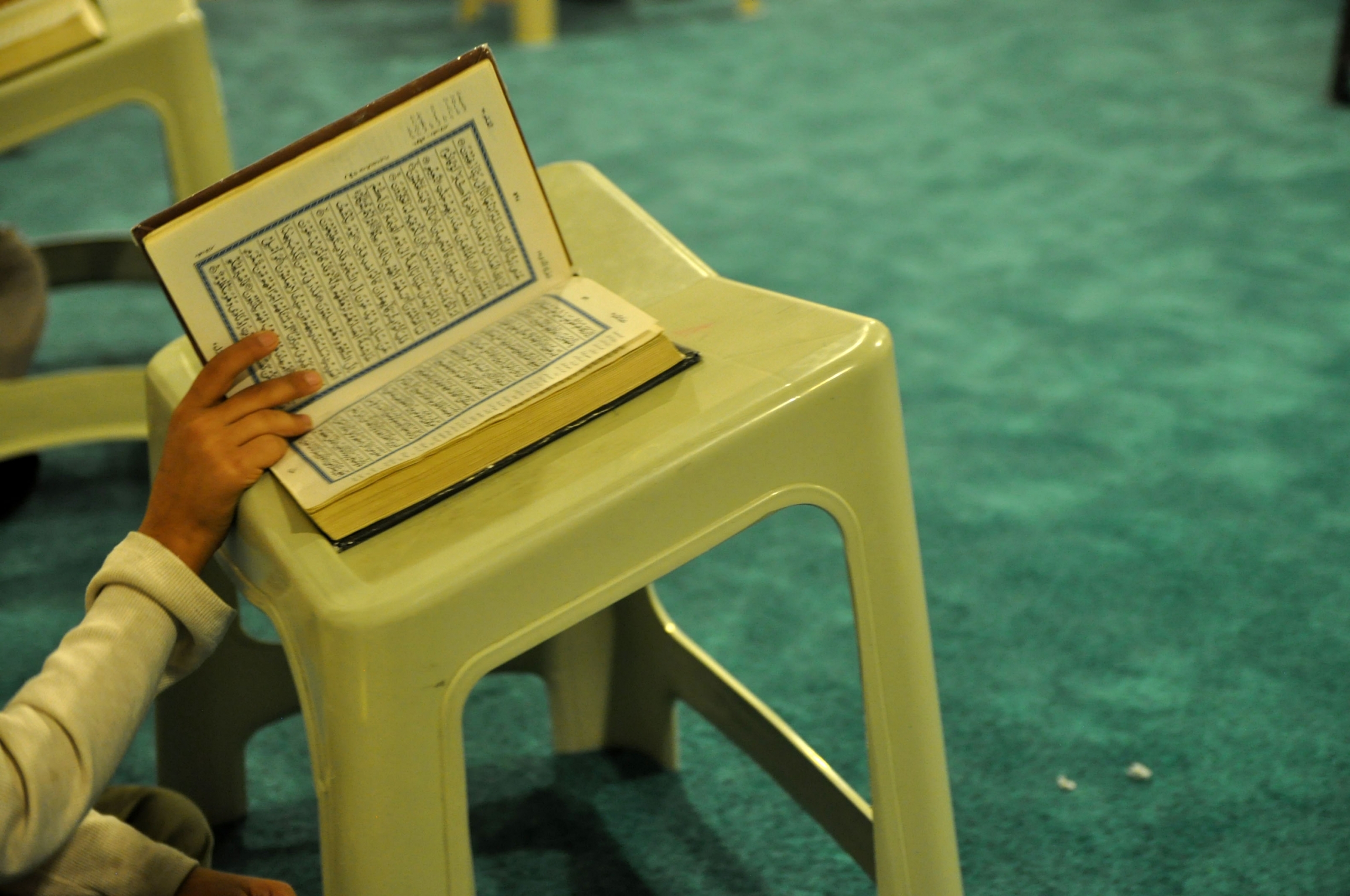 A student recites from the Quran during an evening religious class. In China’s Xinjiang region, the open practice of Islam is often used as grounds to place citizens in "reeducation camps." A hand holds the Quran open on a stool near green carpet