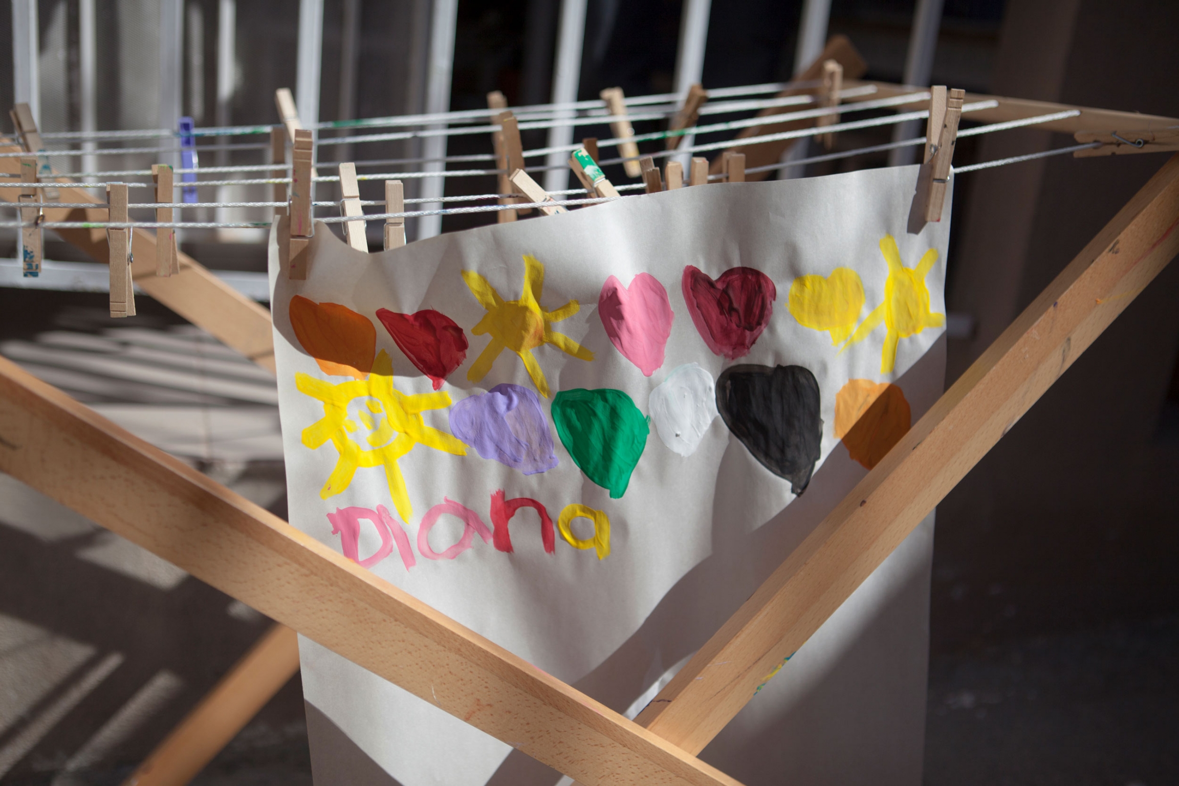 Paintings dry on a rack in the outdoor art area at the Nest. A white paper is shown with colorful hearts painted on it hanging from a wooden rack.