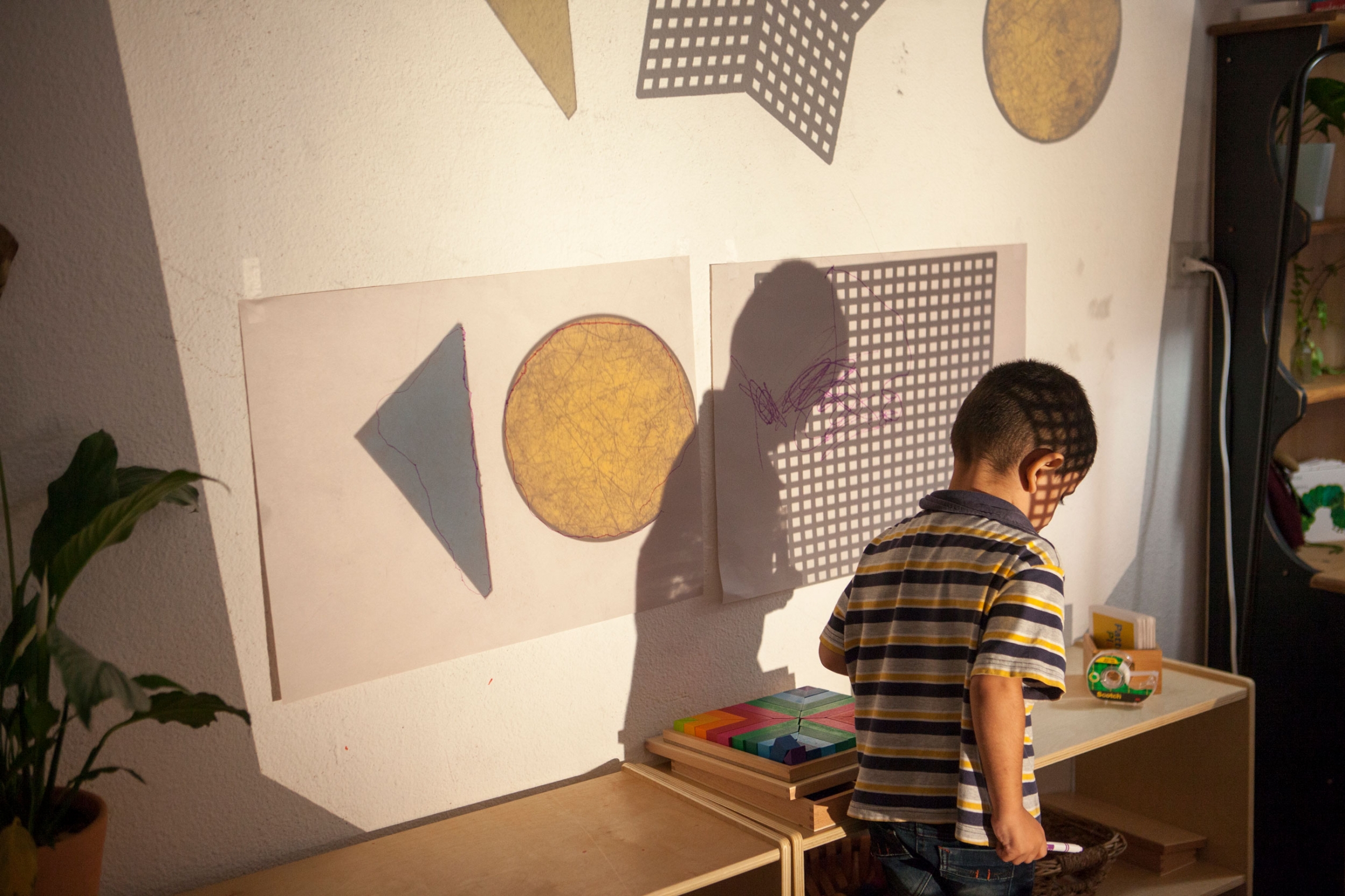 A child walks through shapes projected on a wall at the Tijuana Nest. A child is shown walking past a table with wooden toys.