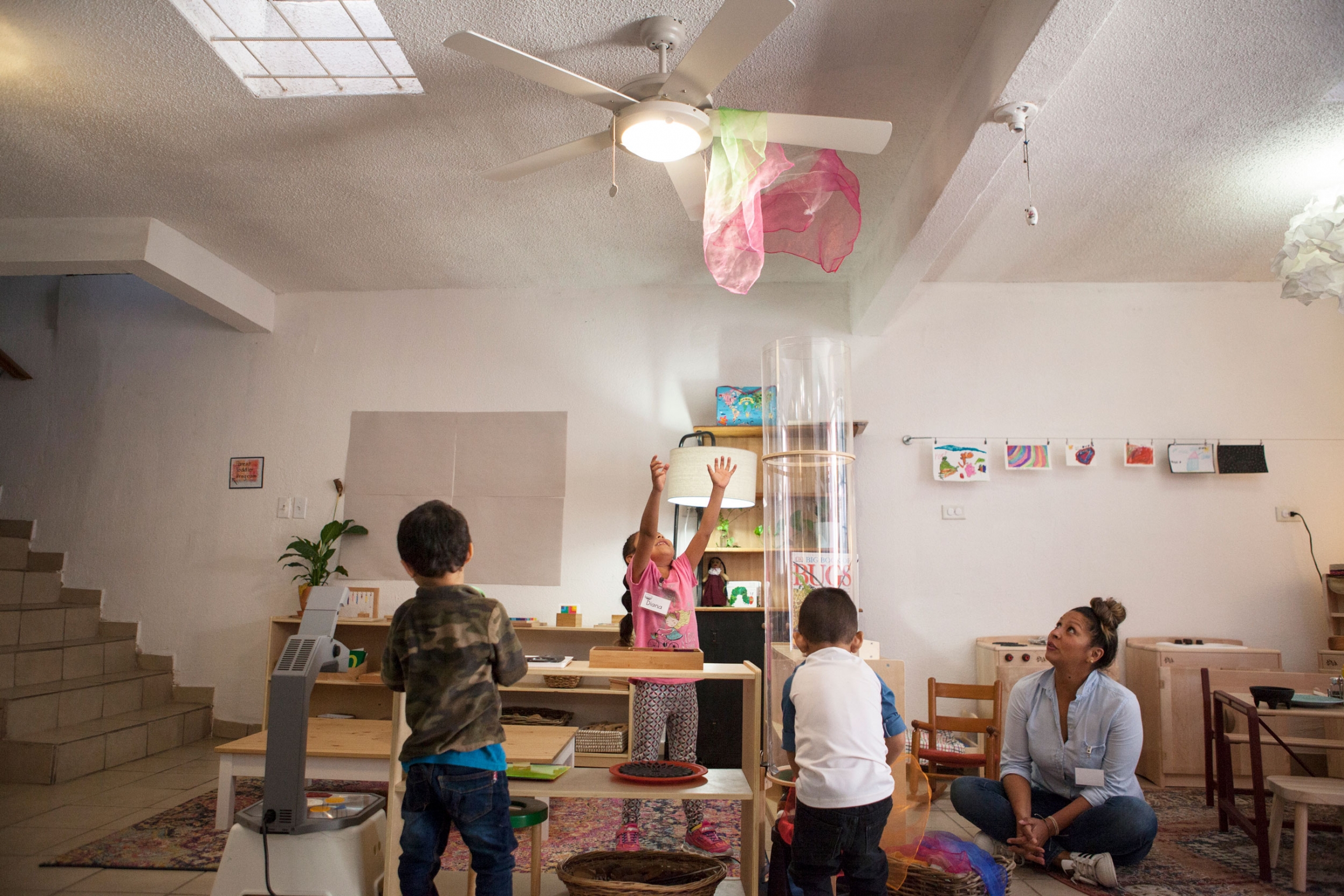 Children play while a scarf gets caught in a ceiling fan. Several children are shown playing with a pink and white scarf shown caught attached to a ceiling fan.
