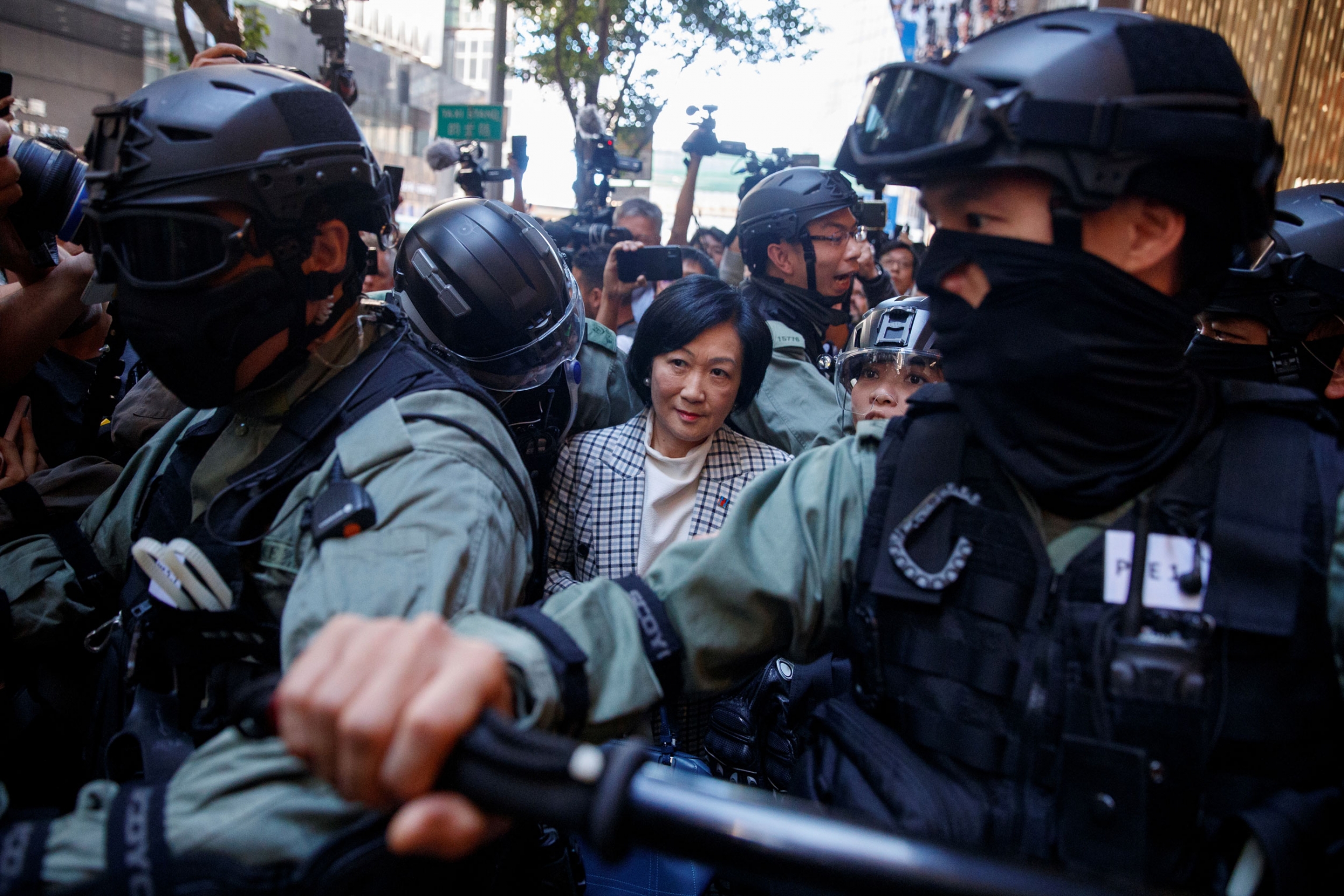 Police escort senior pro-establishment lawmaker Regina Ip after she turned up at an anti-government protest in the Central district of Hong Kong, Nov. 25, 2019. A woman is shown wearing a checkered suit coat and surrounded by an armed police escort.