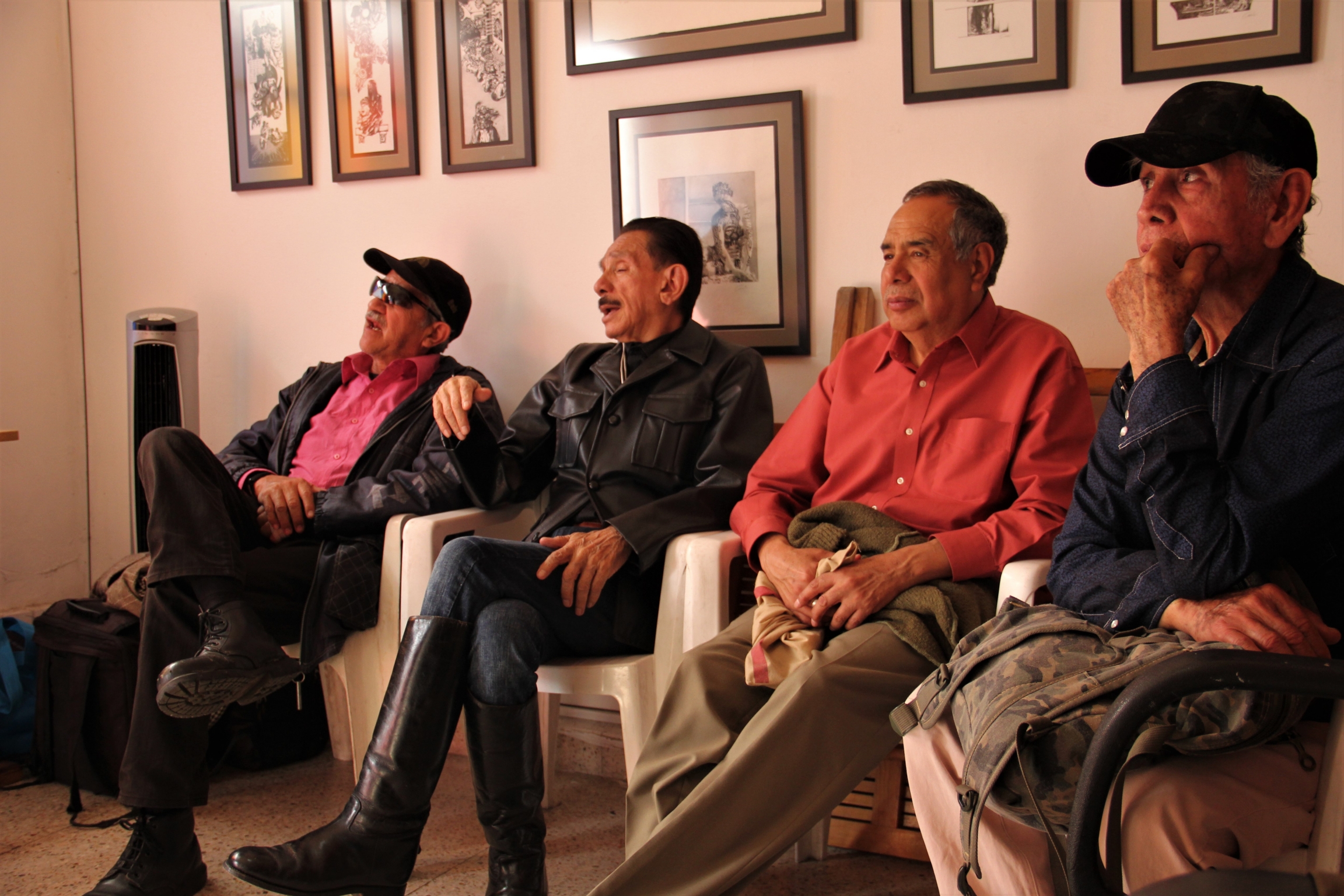Gustavo Pérez, dressed in a black leather jacket and boots, chats with other Vida Alegre members. Four men sit on chairs in a small room