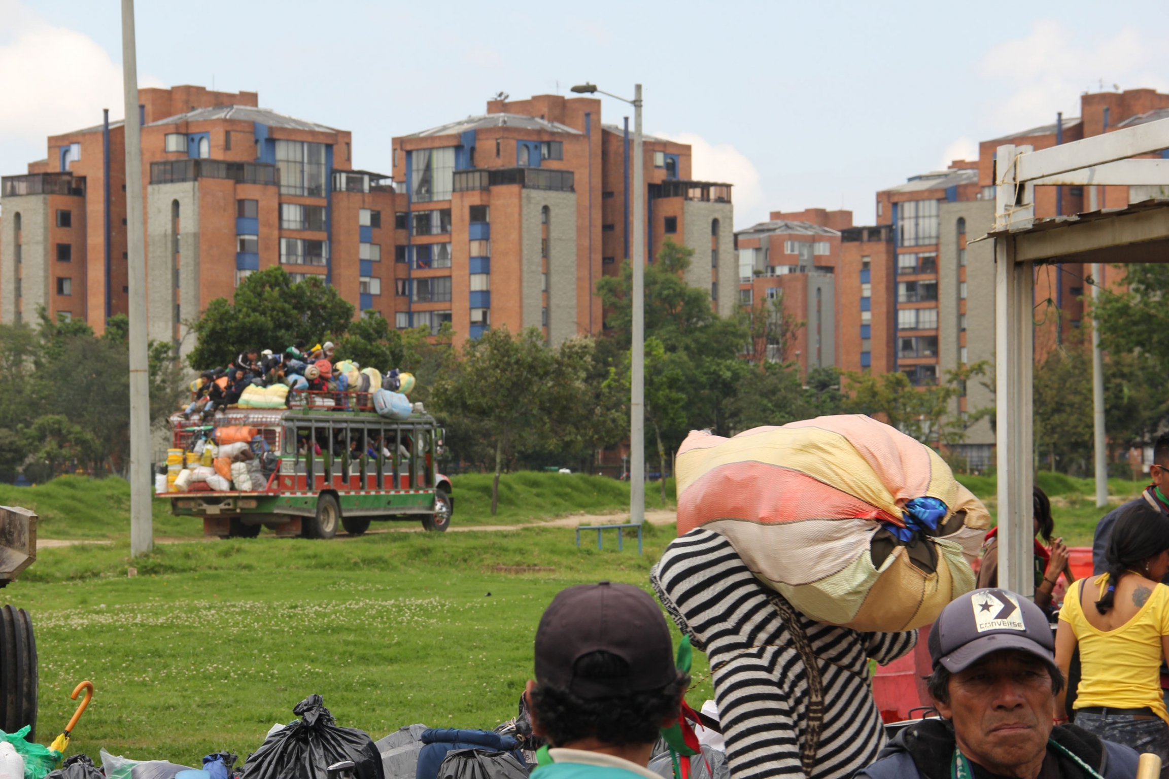 After hours of packing, one of the final chivas sets off on the 15 hour ride back to Cauca. People are crammed inside and even sit on the roof for the arduous journey. A colorful bus is shown in the distance driving away.