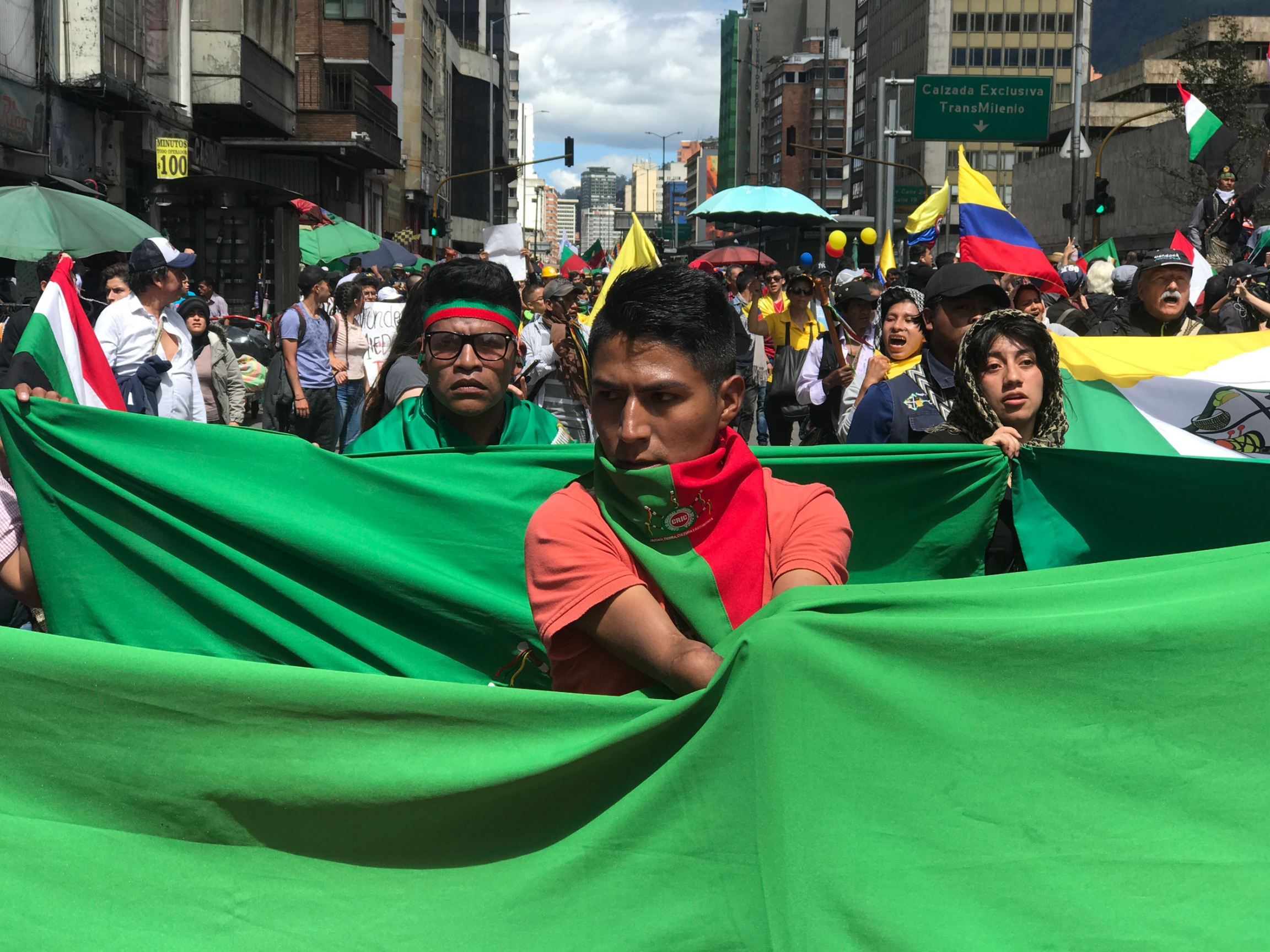 An Indigenous man who lost both arms in an electrical accident as a child carries the red and green banner of the Regional Indigenous Council of Cauca along the streets in downtown Bogota during the most recent national strike on December 4. A man is shown wearing a green and red scarf and carrying a large green flag.
