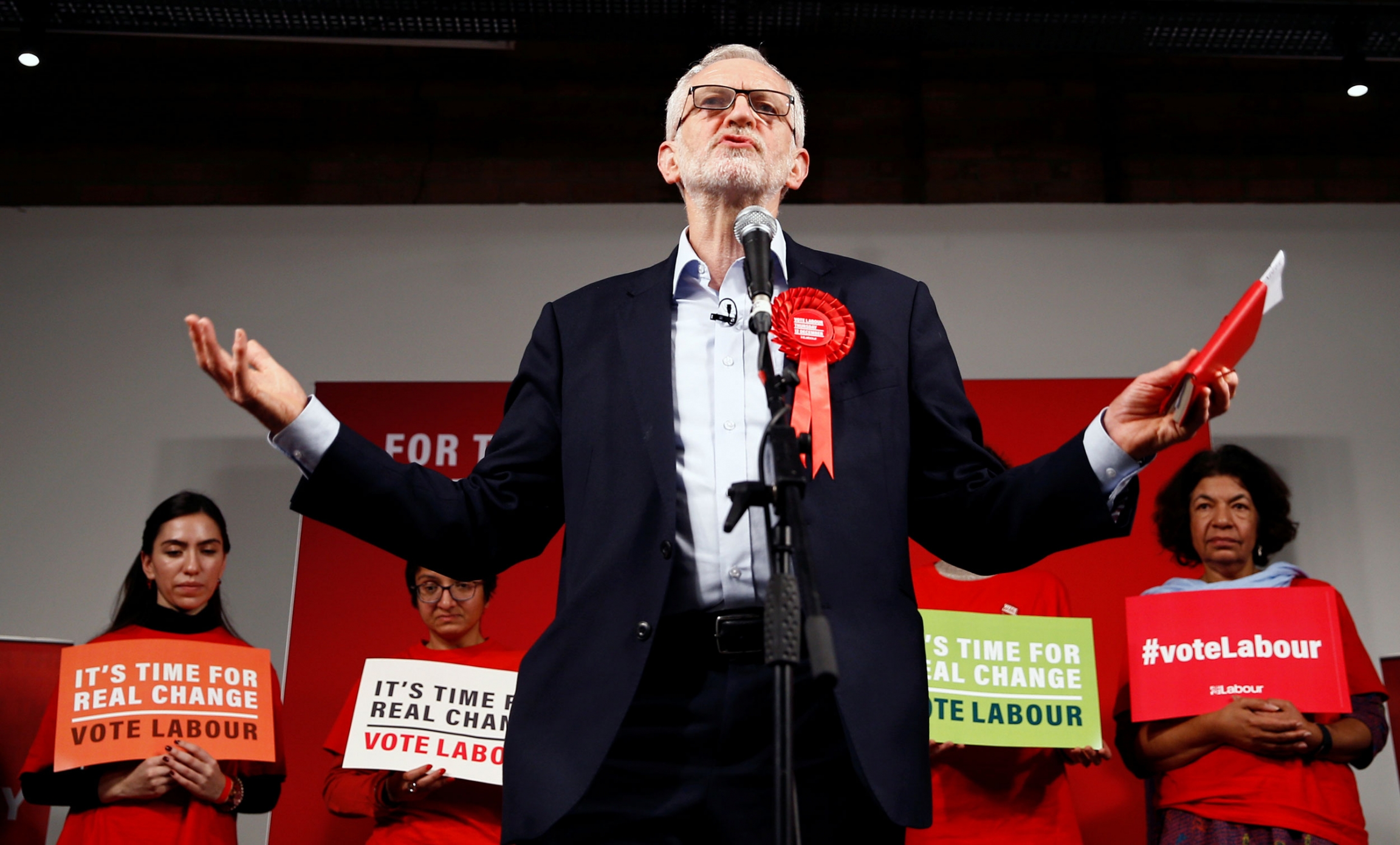 Britain's opposition Labour Party leader Jeremy Corbyn speaks during a final general election campaign event in London, Britain, Dec. 11, 2019. Britain's opposition Labour Party leader Jeremy Corbyn is shown standing a microphone wearing a large red ribbon and holding a red book.
