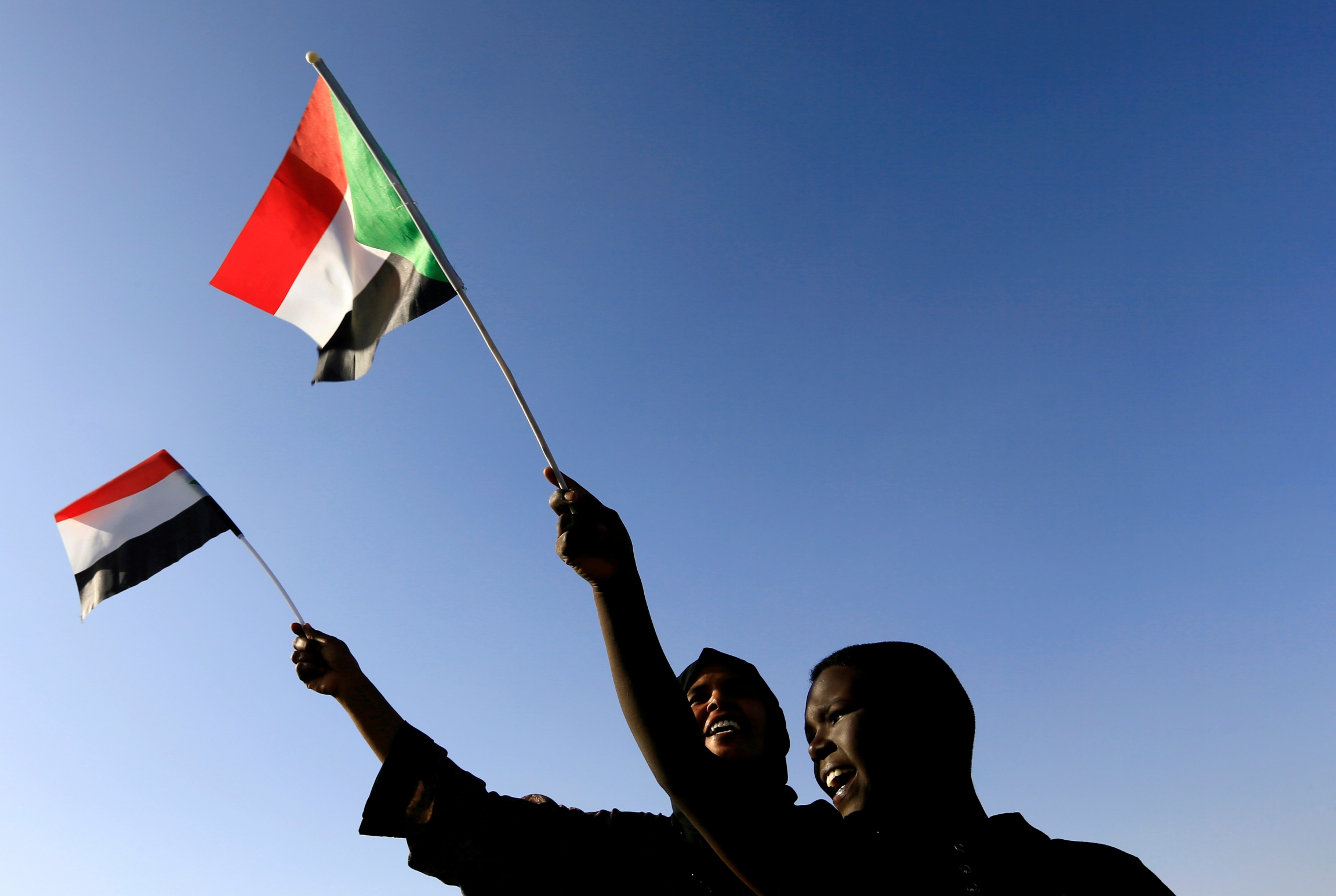 Sudanese civilians wave their national flag as they gather at the freedom square during the first anniversary of the start of the uprising that toppled long-time ruler Omar al-Bashir. Two Sudanese people wave their flag against blue backdrop