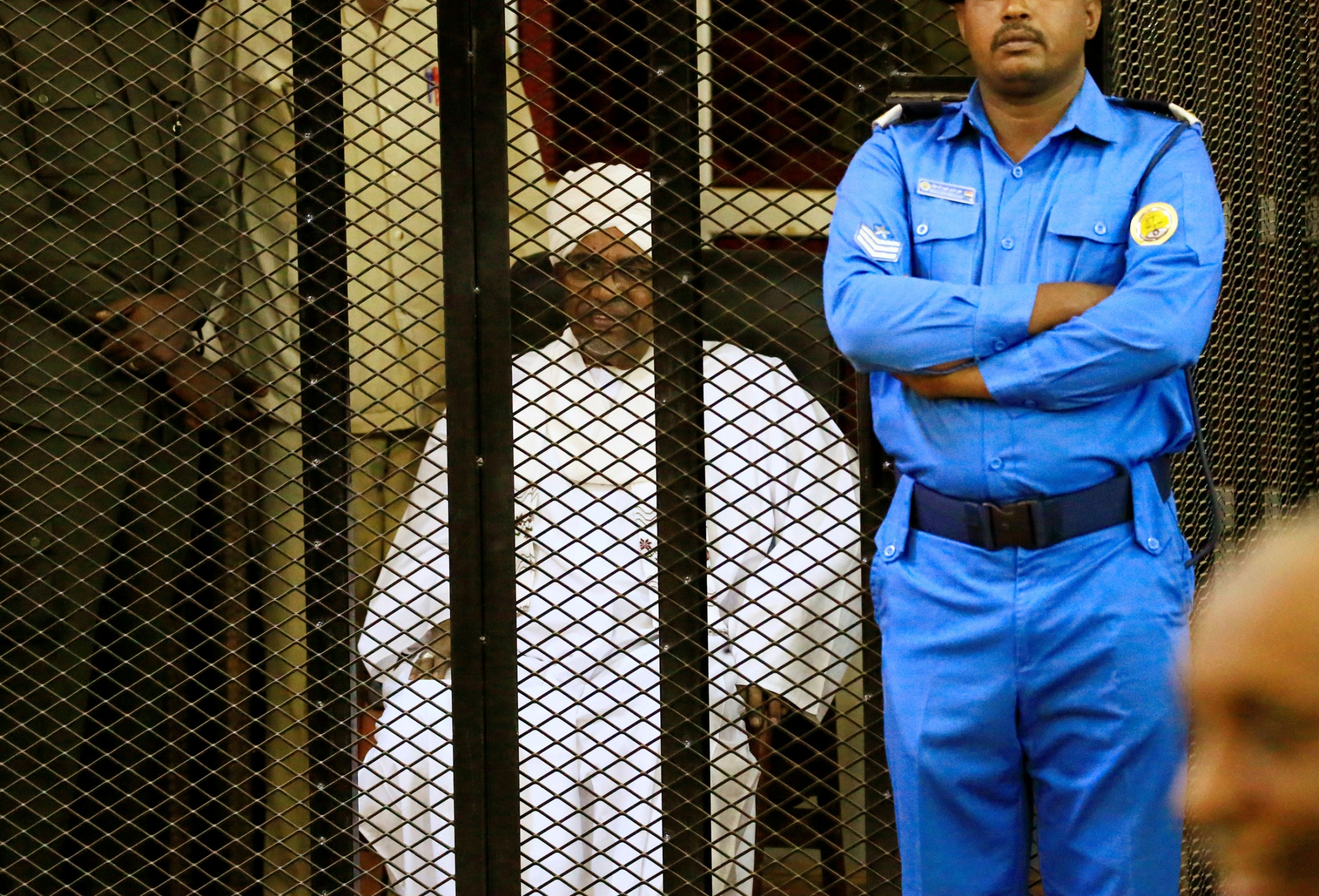 Sudanese former president Omar al-Bashir sits inside a cage during the hearing of the verdict that convicted him of corruption charges in a court in Khartoum, Sudan, Dec. 14, 2019. Sudanese former president Omar al-Bashir is shown sitting in all white and inside a metal cage with a guard standing beside it.