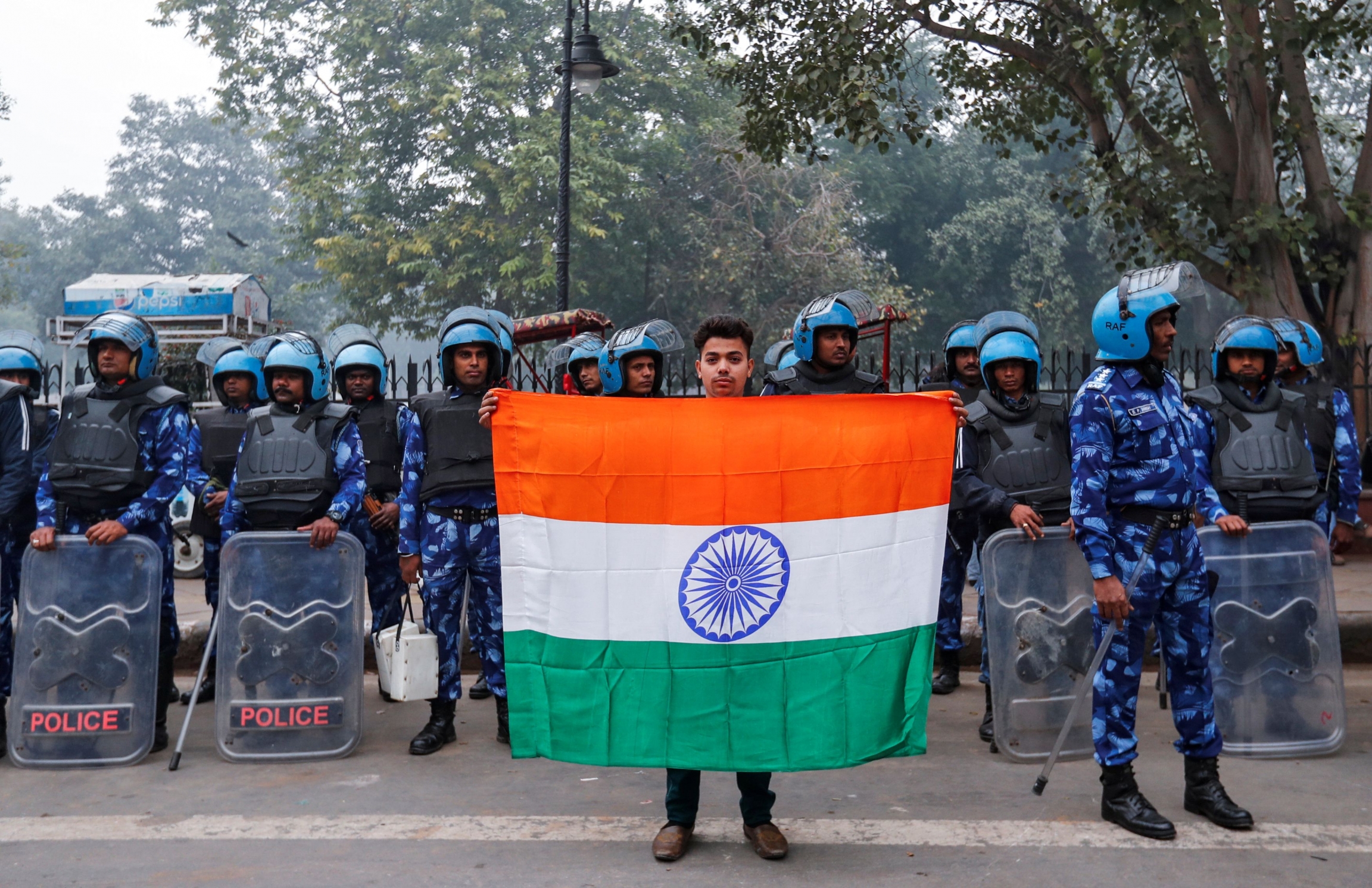 Mohammad Anas Qureshi, 20, who is a fruit vendor, poses for photo with the national flag of India in front of riot police during a protest against a new citizenship law in Delhi, India, Dec. 19, 2019. A man holds Indian flag in front of police wearing blue uniforms.