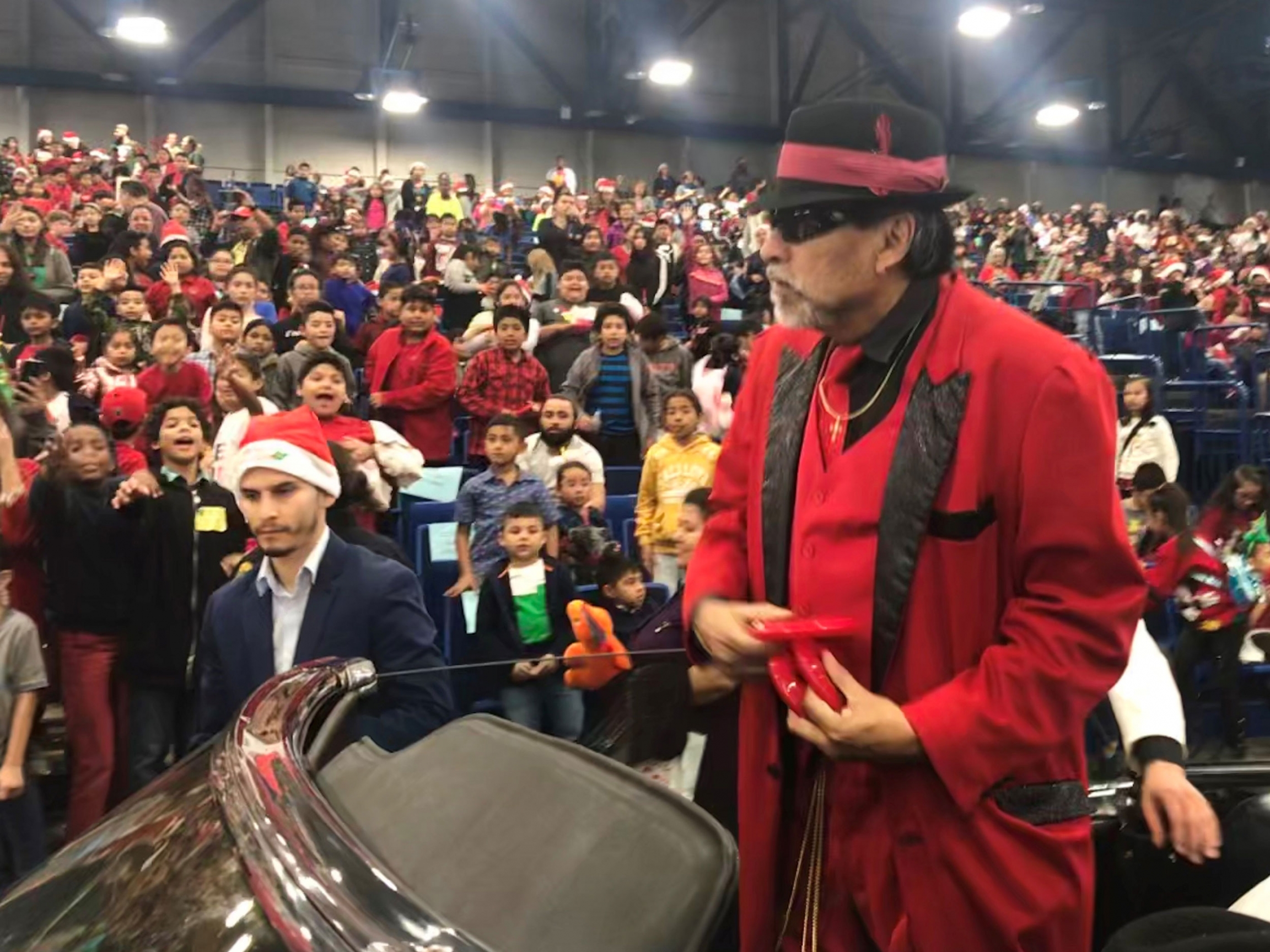 Pancho Claus has been performing at Navidad en el Barrio for more than 30 years. Pancho Claus is shown in a red jacket and black hat standing in a car with a large crowd behind him.