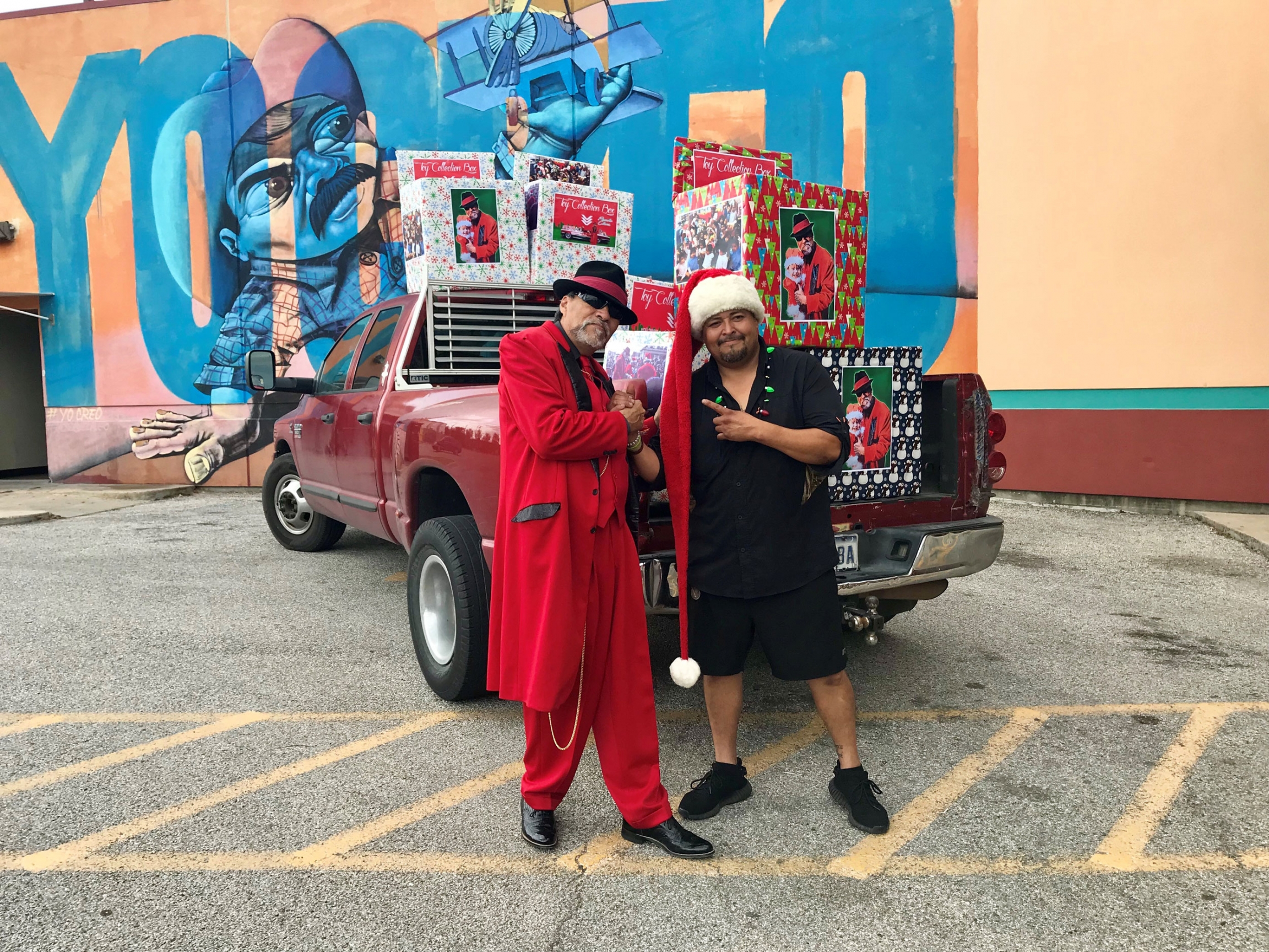 Richard Reyes poses with a volunteer who is dropping off toy collection boxes to different locations around town. Richard Reyes is shown wearing a long red coat and pants and standing in front of a pickup truck.