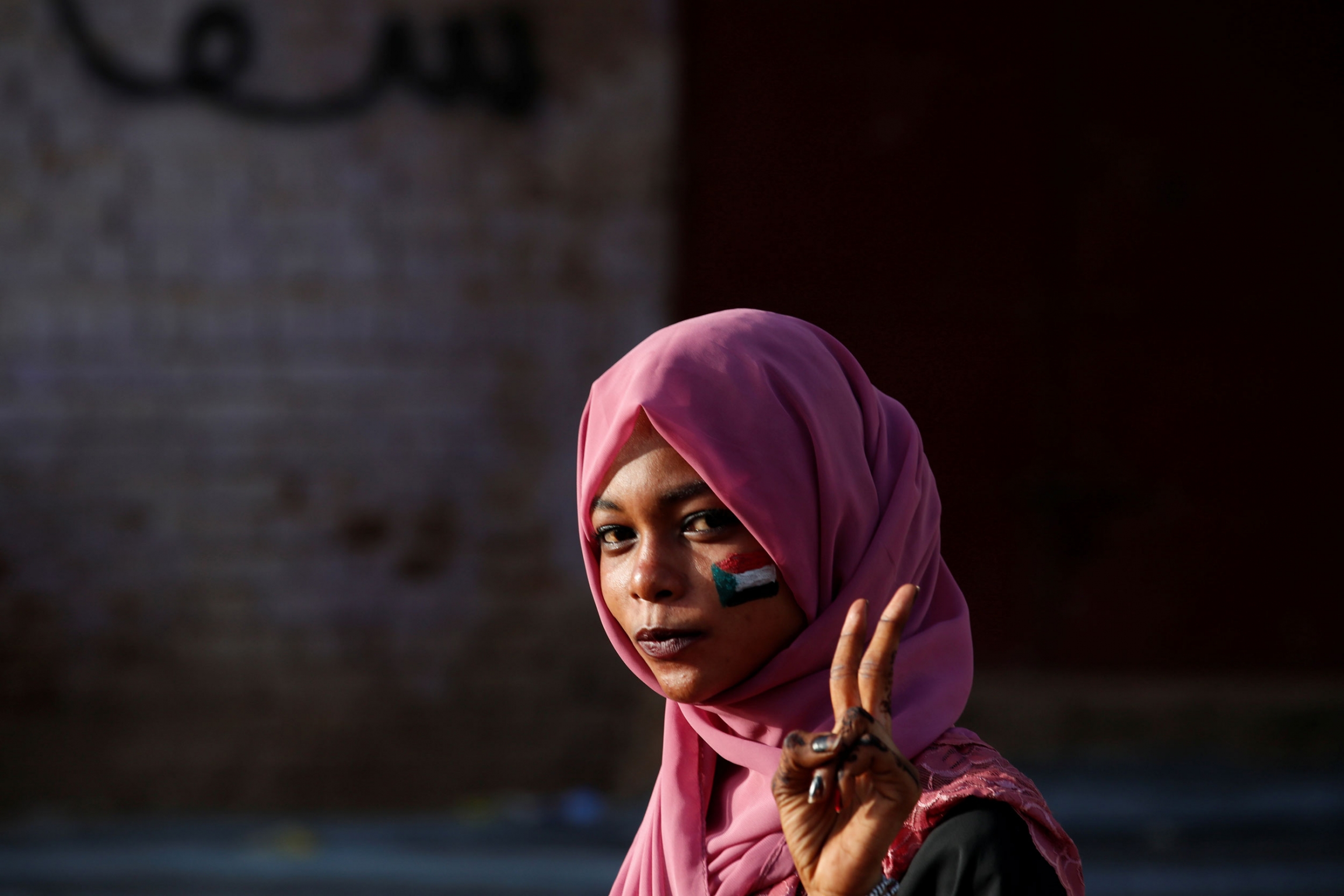 A Sudanese demonstrator makes victory sign as she protests outside Defense Ministry in Khartoum, Sudan, April 16, 2019. A woman is shown wearing a pink headscarf and with a Sudanese flag painted on her cheek.