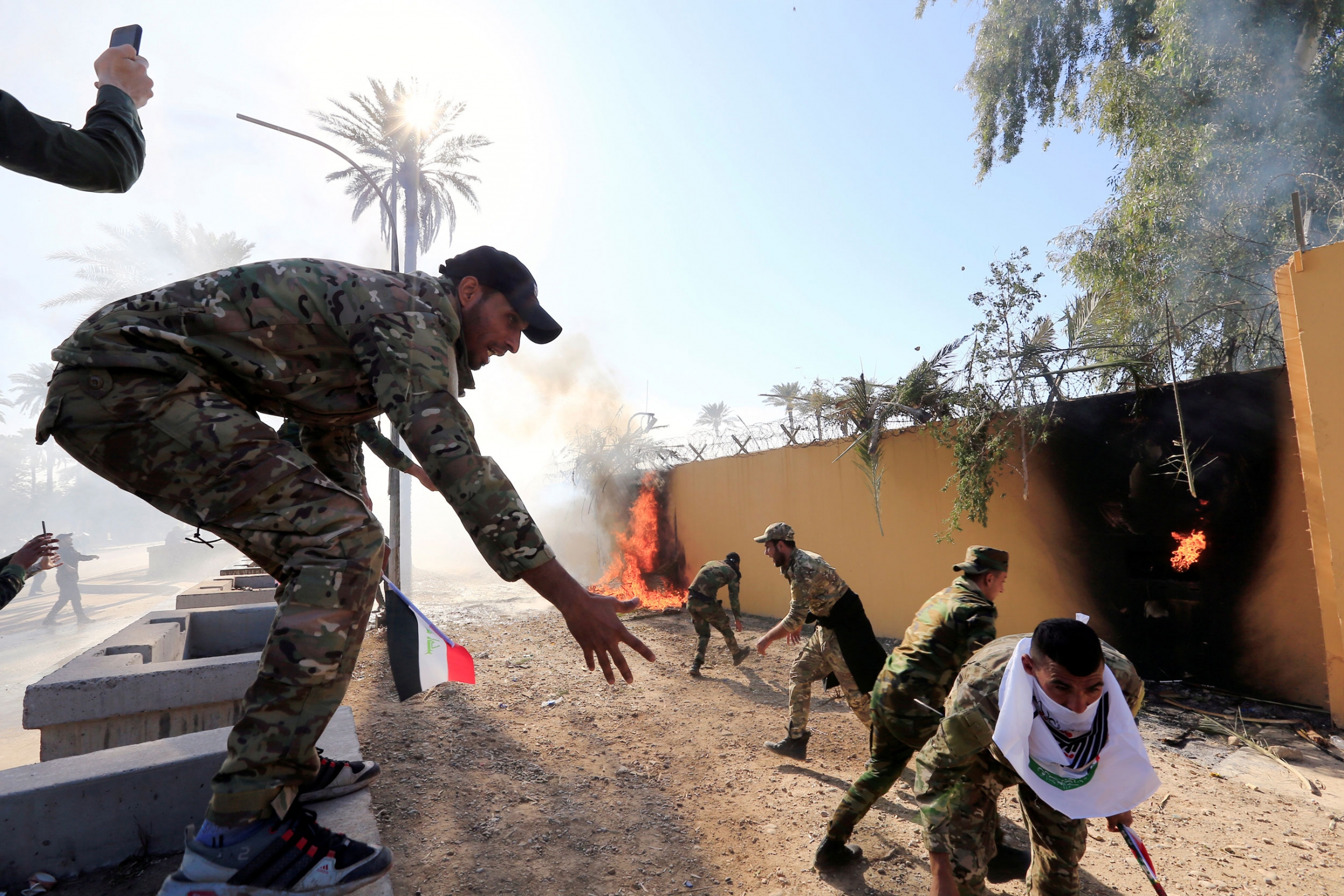 Hashd al-Shaabi paramilitary forces fighters set fire on the US Embassy wall to condemn air strikes on their bases, in Baghdad, Iraq, Dec. 31, 2019. Several men are shown wearing military fatigues outside of a khaki-colored wall that has been set on fire.