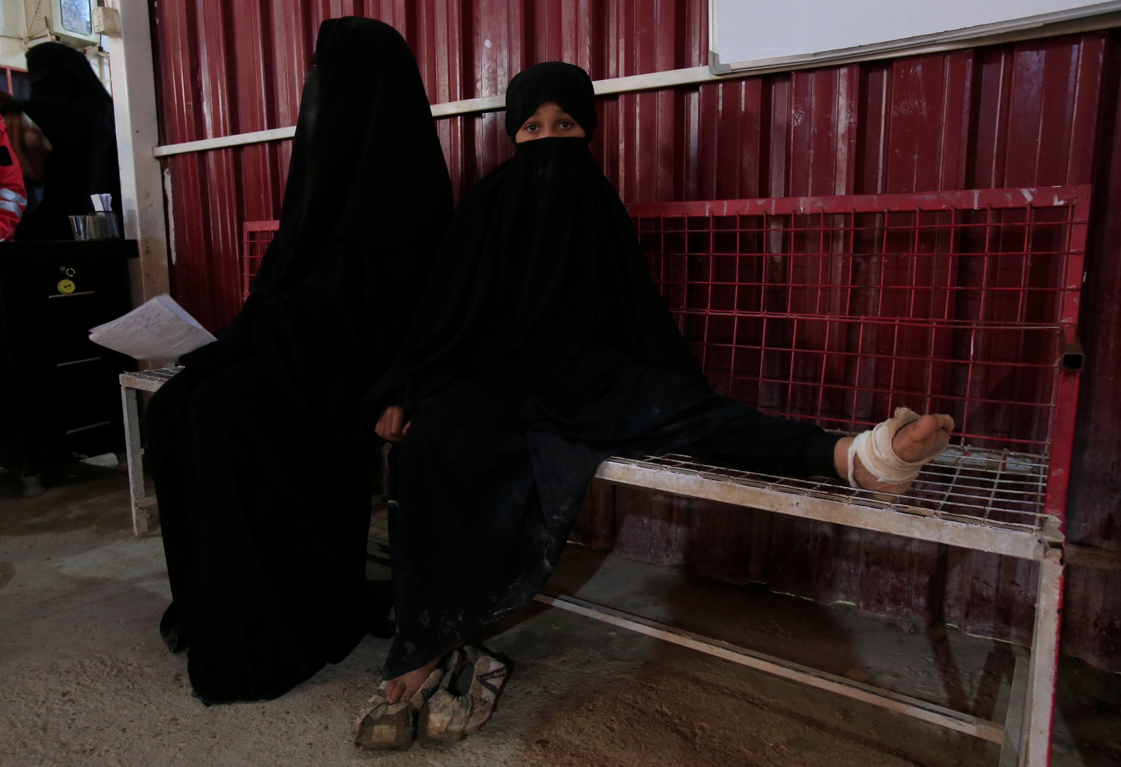 A wounded girl sits at a clinic at al-Hol displacement camp in Hasaka governorate, Syria, April 2, 2019. Two girls wearing black clothing sit on a bench in a clinic, one has a wounded foot wrapped in bandage