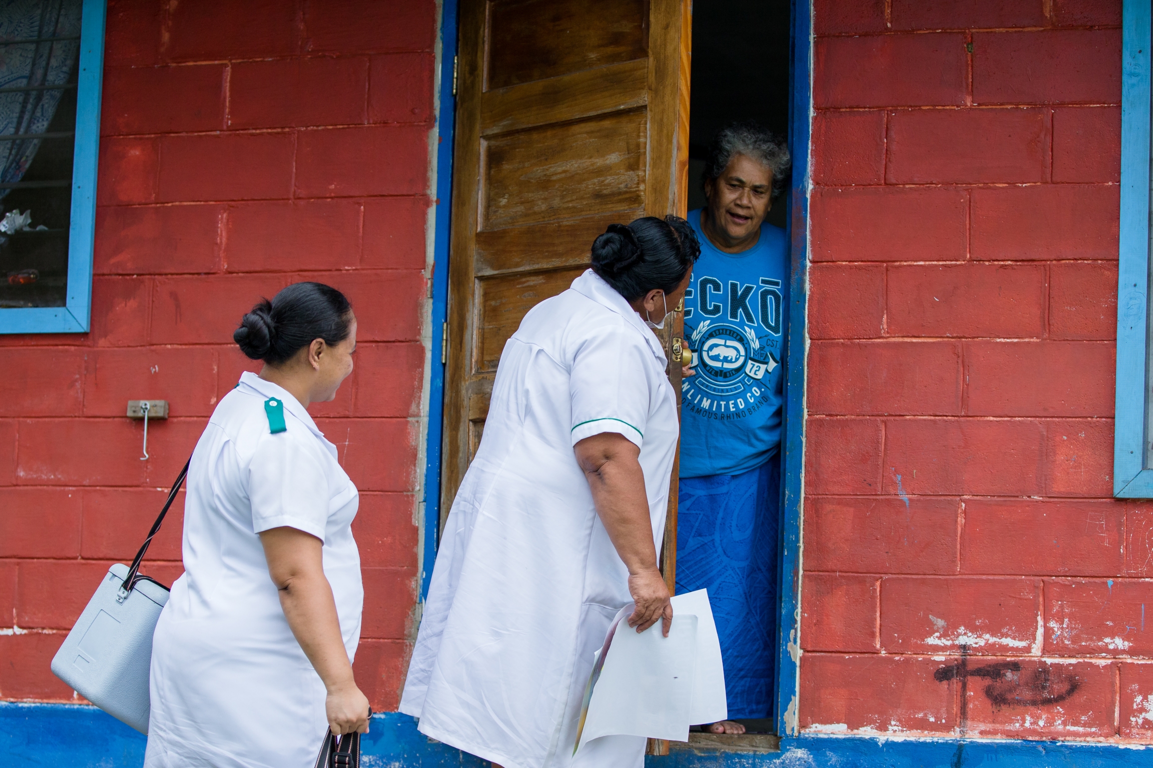 On Dec. 2, 2019, nurses visit homes in Satuimalufilufi village during a UNICEF-supported national vaccination campaign in response to the current measles outbreak in the Pacific region. Two nurses stand in front of a house painted red and talk to a person standing at the door