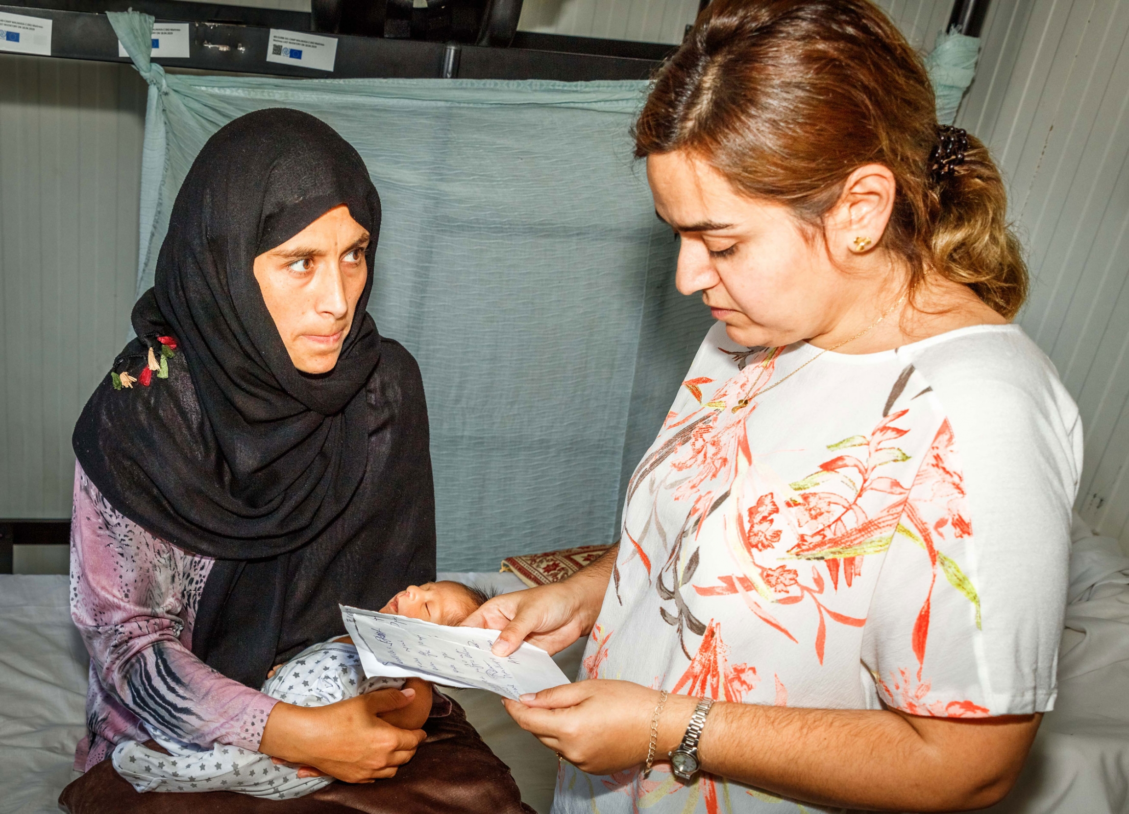 Inside a container in the Malakasa refugee camp in Athens, refugee advocate, Sahar Kamrani (right) looks at paperwork that Nadereh Shahbazi (left) doesn't understand. Kamrani spends her days helping families navigate the complicated asylum process in Gree A woman is shown wearing a dark head scarf while holding an infant and looking at another woman holding papers.