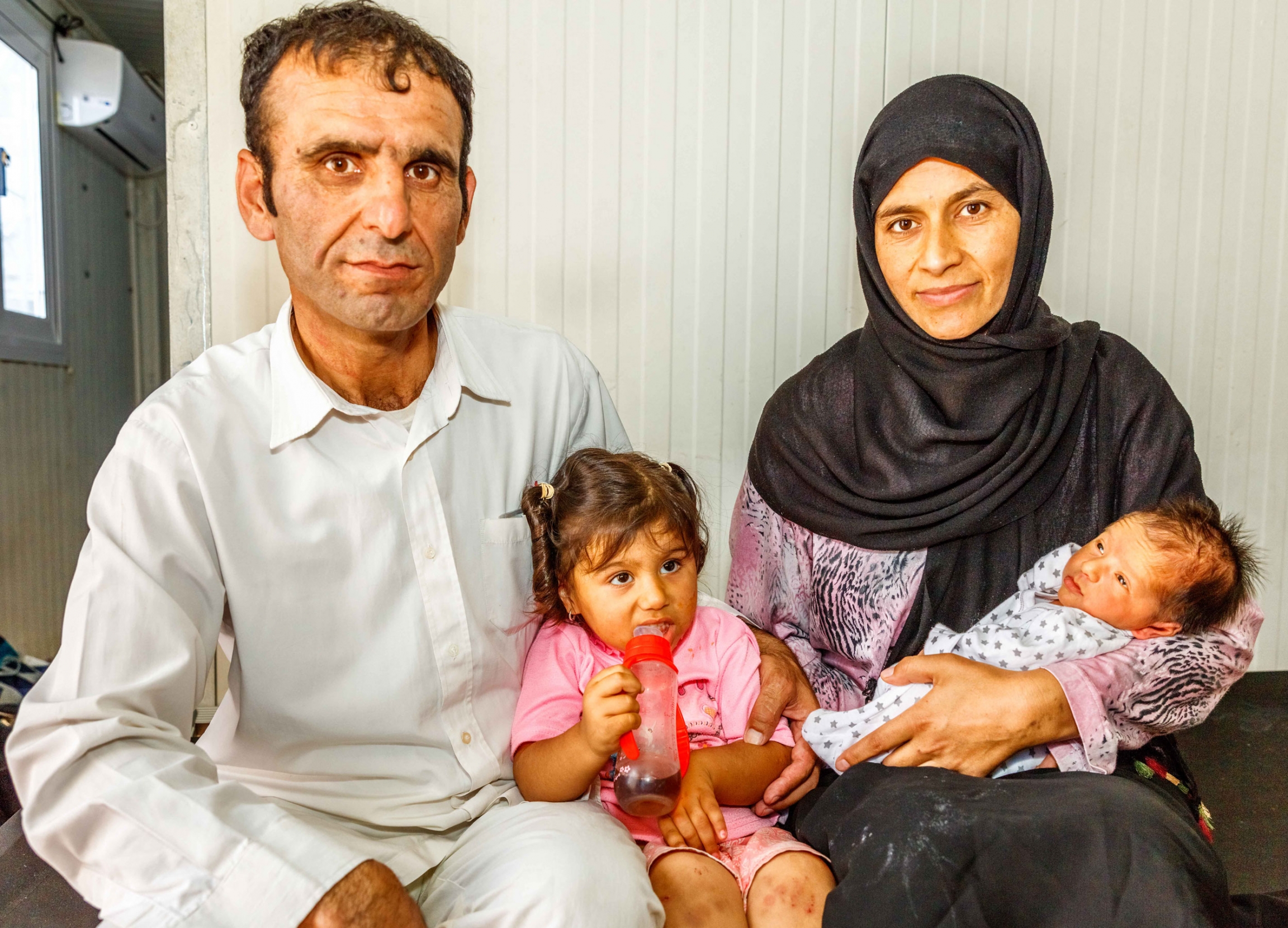 (Left to right) Azimollah Shahbazi, Boshra (aged 2), Nadereh Shahbazi and Baby Mustafa in their container home — given to them after Mustafa was born — by the International Organization of Migration in the Malakasa refugee camp in Athens. A women is shown wearing a dark head scarf holding an infant and sitting next to a man wearing all white and a young girl sitting in-between.