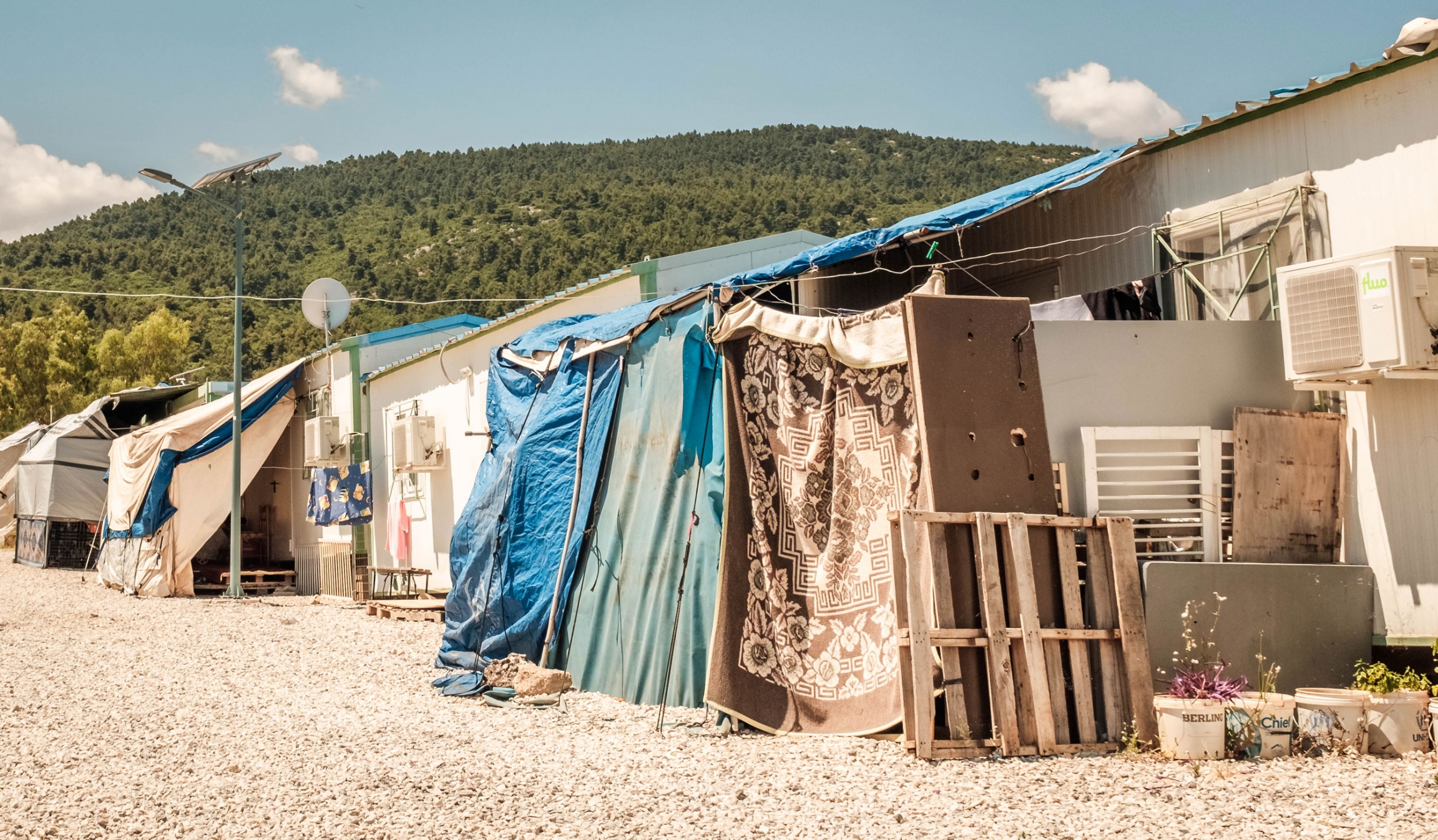 Containers that house asylum seekers in the Malakasa refugee camp are small. Many families construct makeshift shelters adjoining their container for storage of belongings and added space. A row of container homes are shown with additional sheets and blankets attached to the front.