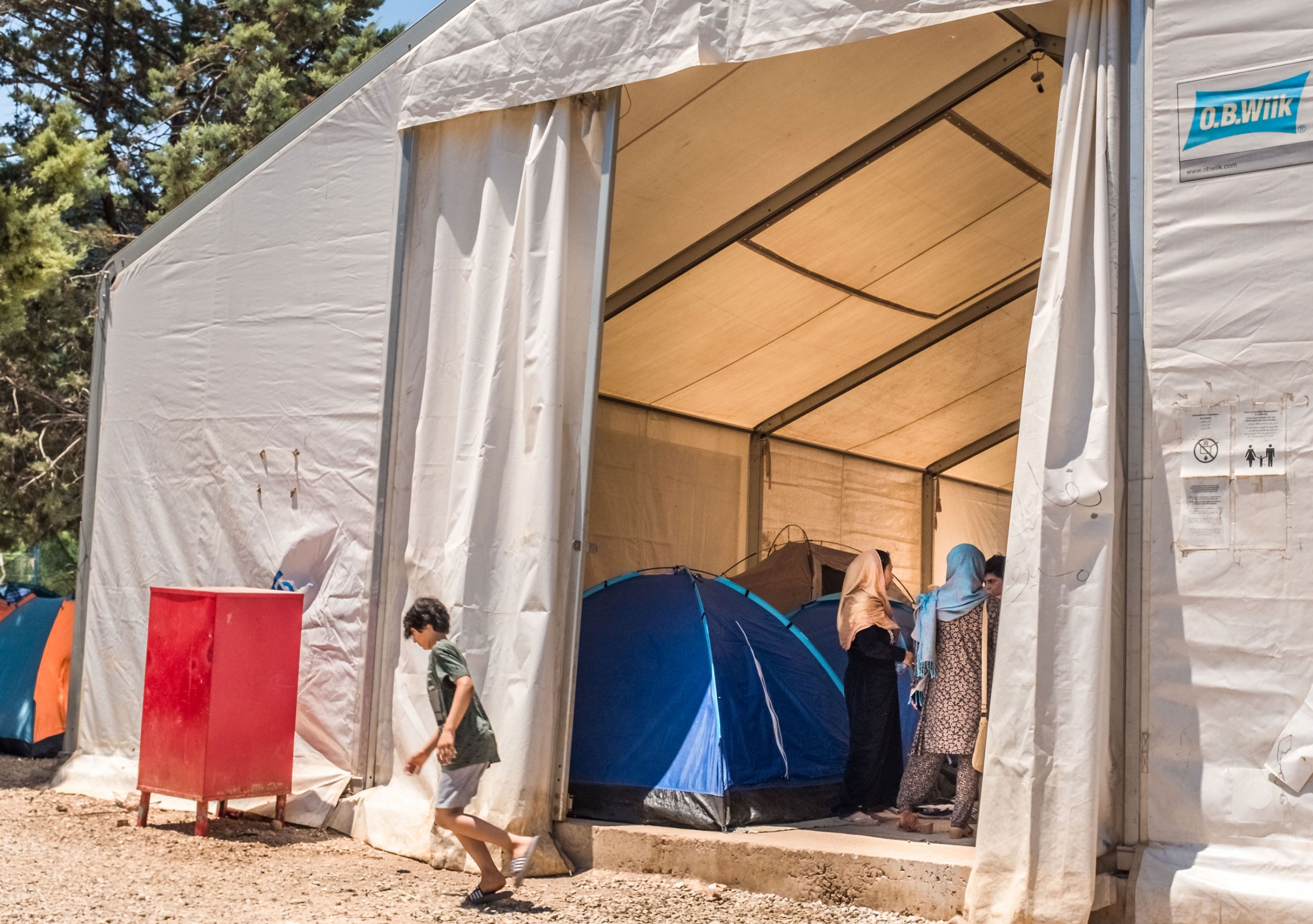There are not enough containers to house the families that arrive almost daily at the Malakasa refugee camp in Athens, so the camp has some bigger tents where asylum seekers can pitch their own tent. A large white tent is shown with a group of women standing among smaller pup tents.