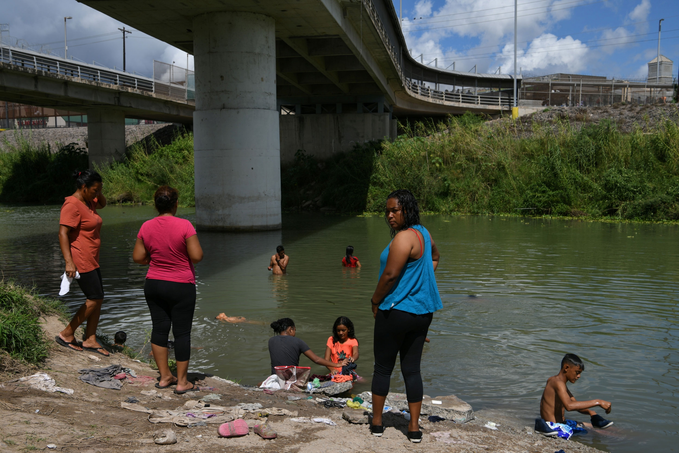 Migrants bathe in the Rio Grande near a makeshift encampment occupied by asylum seekers. Several people are shown standing on the shore and in the Rio Grande river under a large bridge.