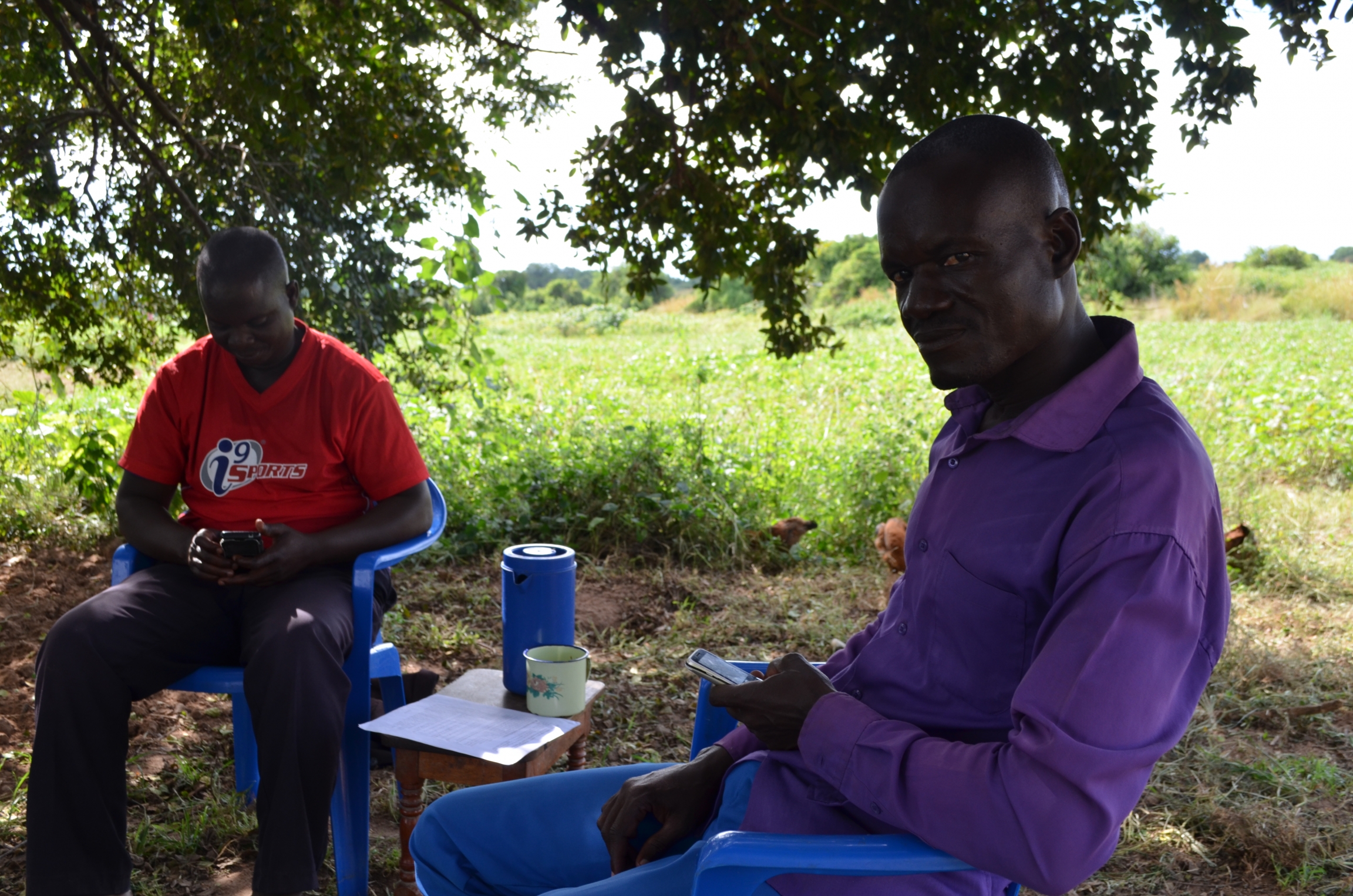 Fred Mwesigwa, 45, (right) sits with his brother Moses (left). They say none of their family members has been compensated for the land they were forced off over two years ago. Nov. 30, 2019, Buliisa district, Uganda. A man wears a purple button-down shirt and sits on a plastic chair next to man who wearing a red shirt.