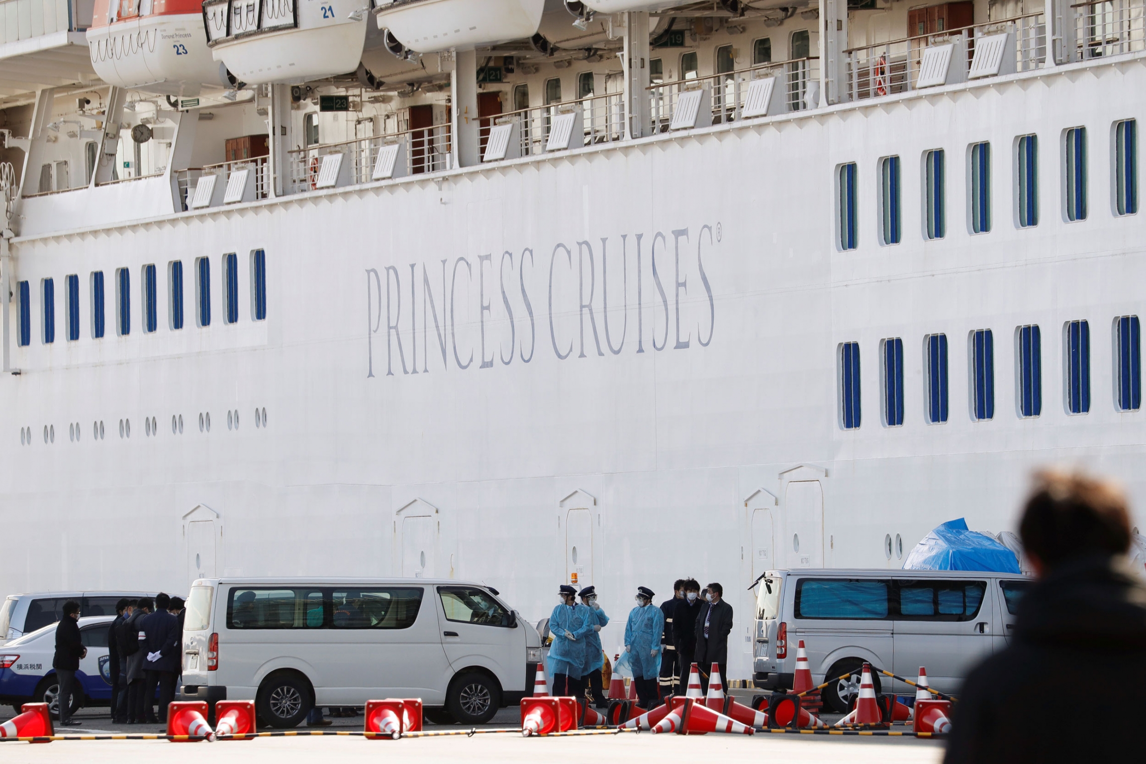 Personnel wearing masks stand near the cruise ship Diamond Princess, where 10 more passengers tested positive for coronavirus, after it arrived at Daikoku Pier Cruise Terminal in Yokohama, south of Tokyo, Japan, Feb. 6, 2020. Several vans are shown parked next to a cruise ship with personnel wearing masks stand nearby,