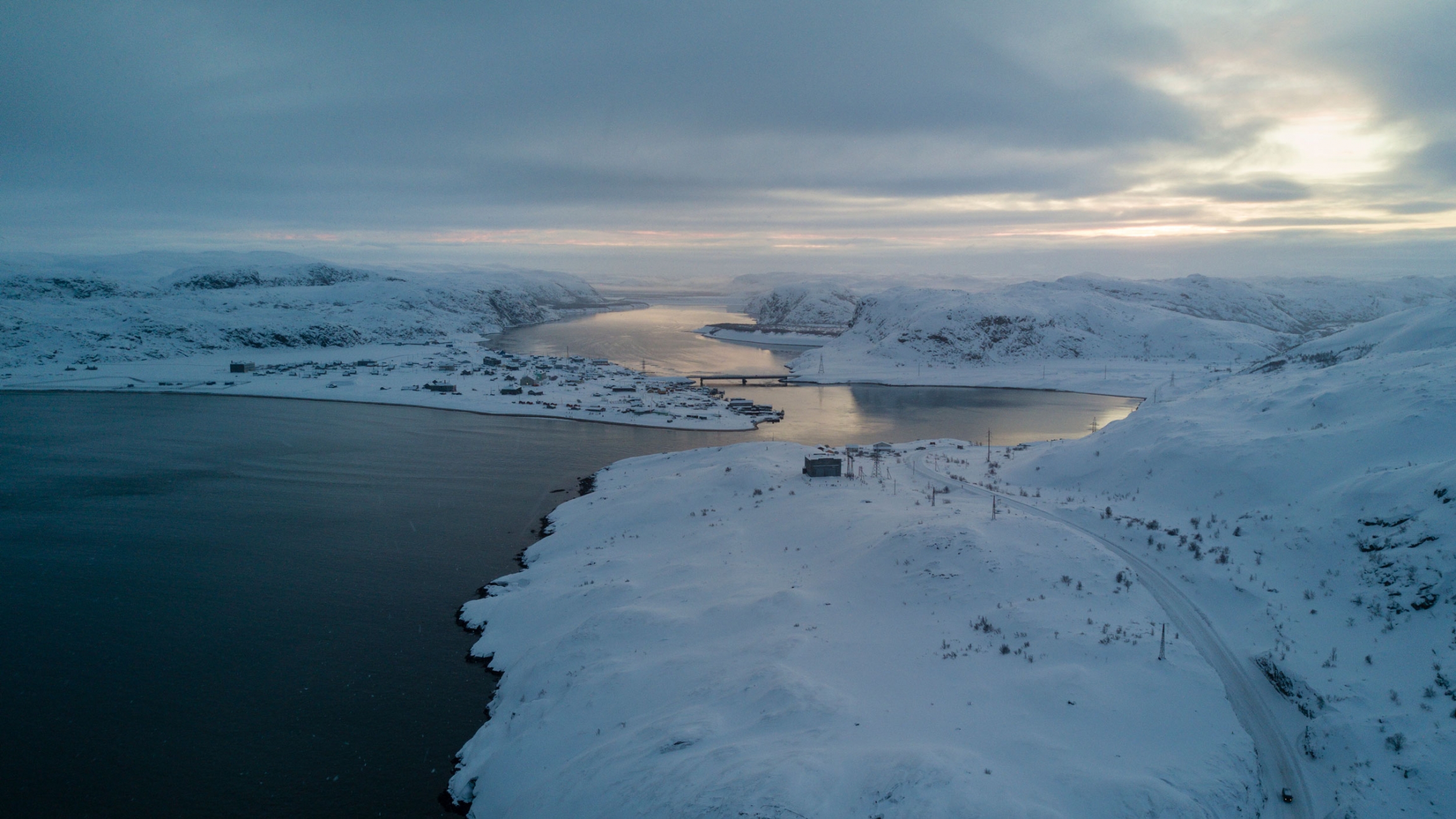 An aerial view of Teriberka village showing a winding blue-colored river and the deep blue color of the Barents Sea coast.