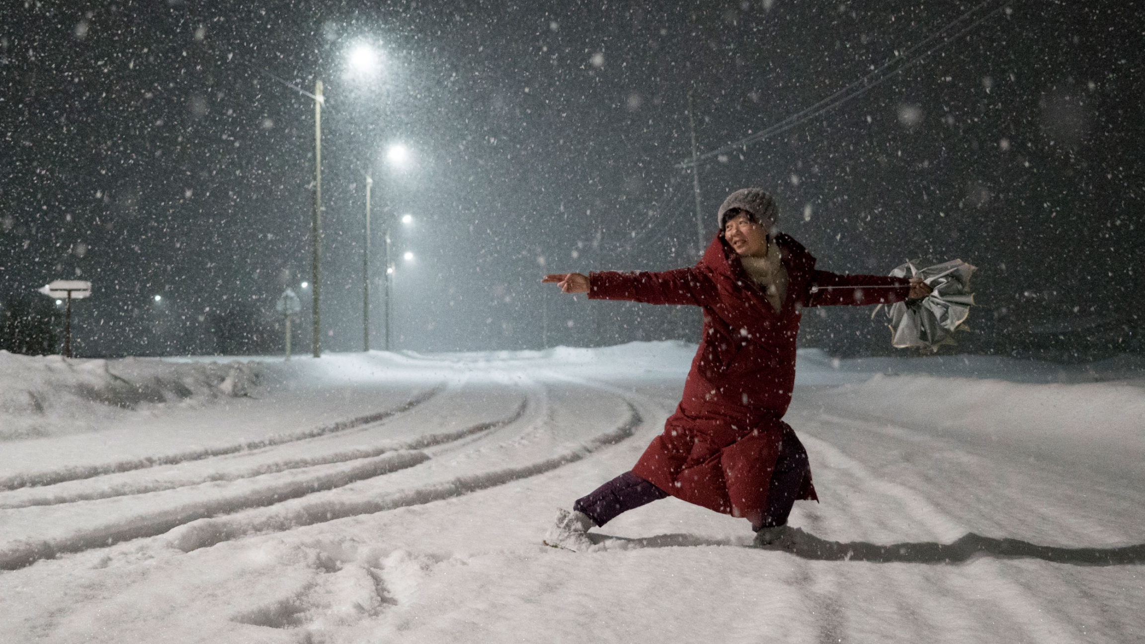A Chinese tourist dances while waiting for bus departure in Teriberka, Russia. A woman in a red jacket is shown dancing in the street with snow falling all around her.