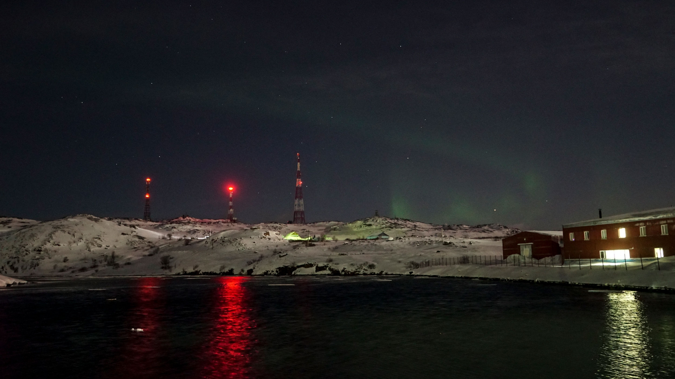 The northern lights at night in Teriberka, Russia. A green swirl of the northern lights is shown against a dark sky with antennas with red lights in the nearground.